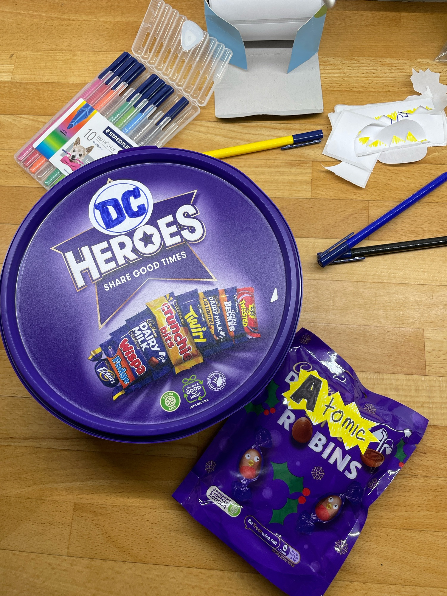 A tub of Cadbury heroes chocolates with a sticker labelling them DC heroes next to a bag of Christmas robin chocolates relabelled Atomic Robins. Felt pens and stickers are in the background. 