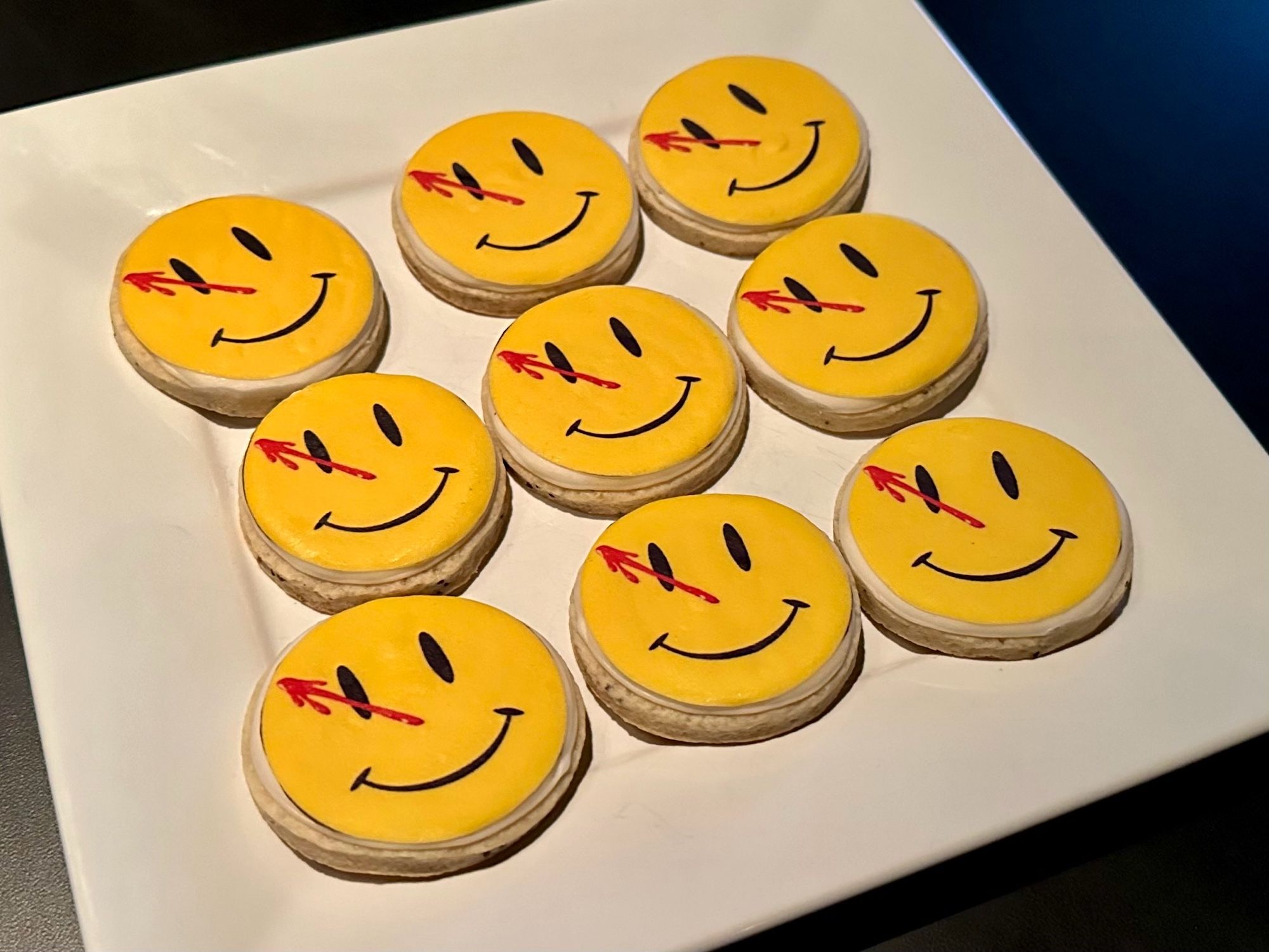 A plate of 9 cookies, each decorated with the iconic Watchmen image of a blood stained, yellow smiley face.