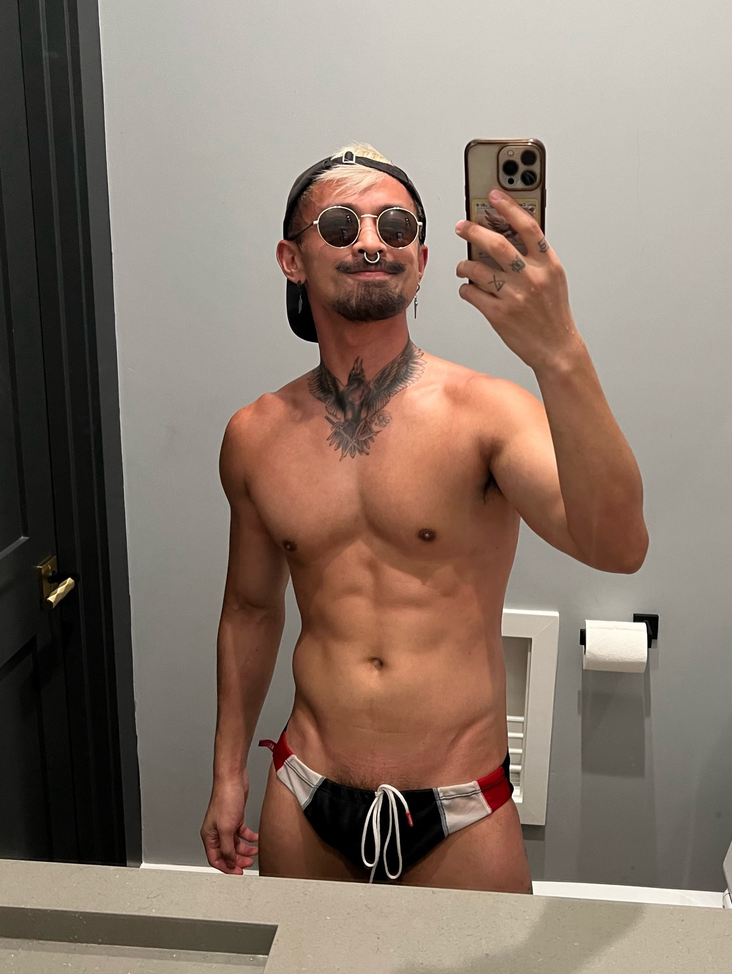 Blonde tattooed bisexual in sunglasses and piercings poses for a mirror selfie. The speedo is barely covering the essential areas