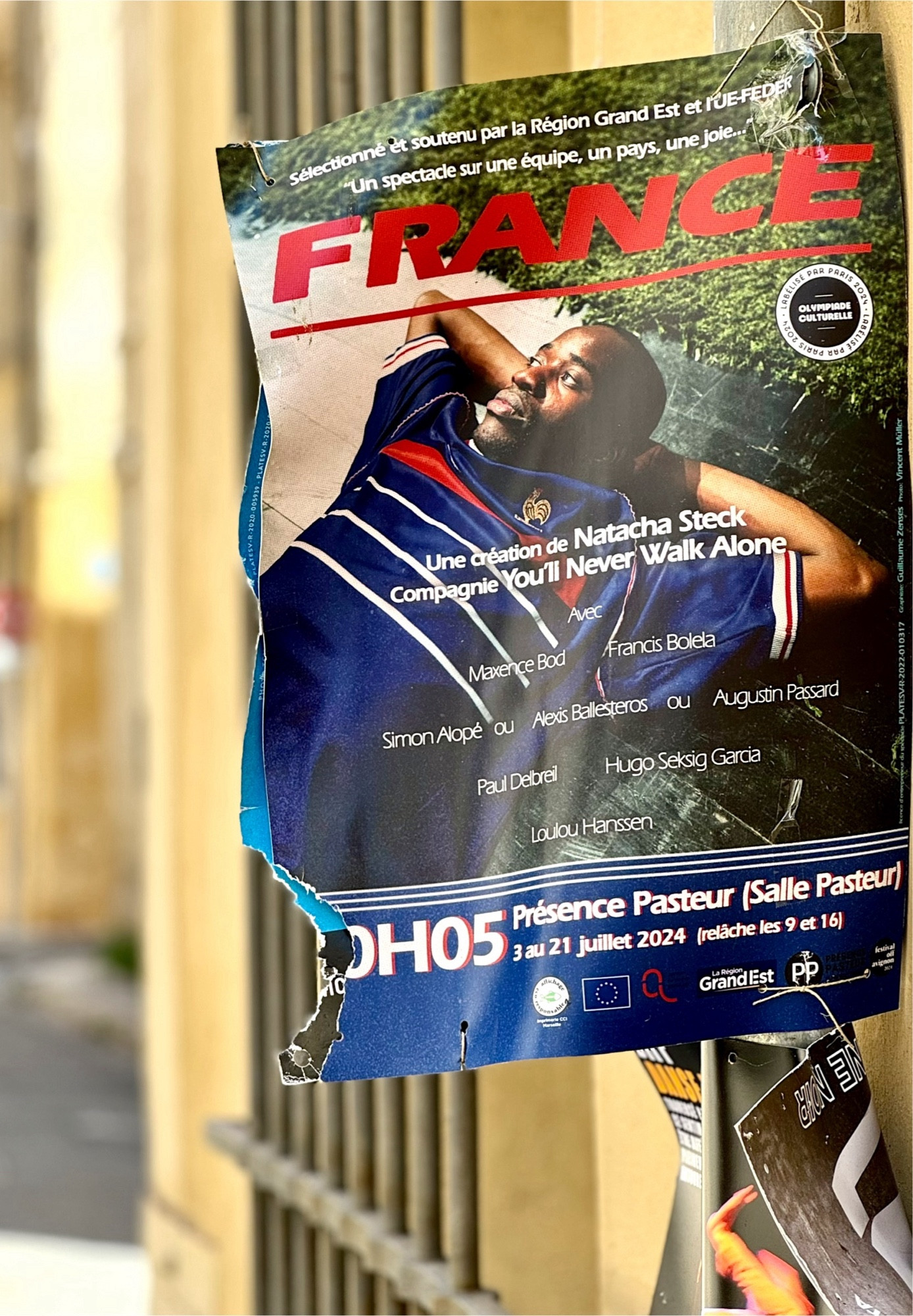 Eine gelbe Hauswand in Avignon. Ein Plakat kündigt eine Theatervorstellung mit dem Titel ‚France‘ an. Ein junger dunkelhäutiger Fußballspieler in dem blauen Nationaltrikot liegt auf dem Rücken und schaut zur Seite