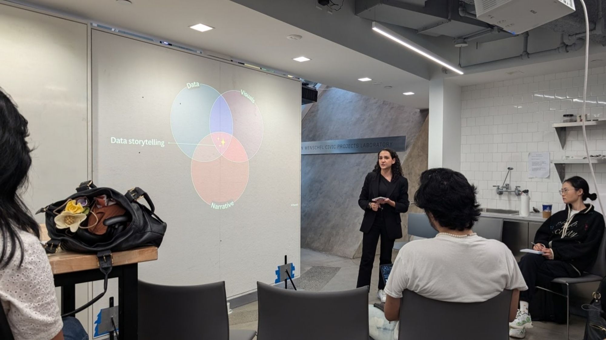 Photo of short female presenter with brown hair standing in front of a slide showing a Venn diagram of three overlapping circles: Data, Narrative, Visuals, and Data storytelling is the middle intersection of all three.