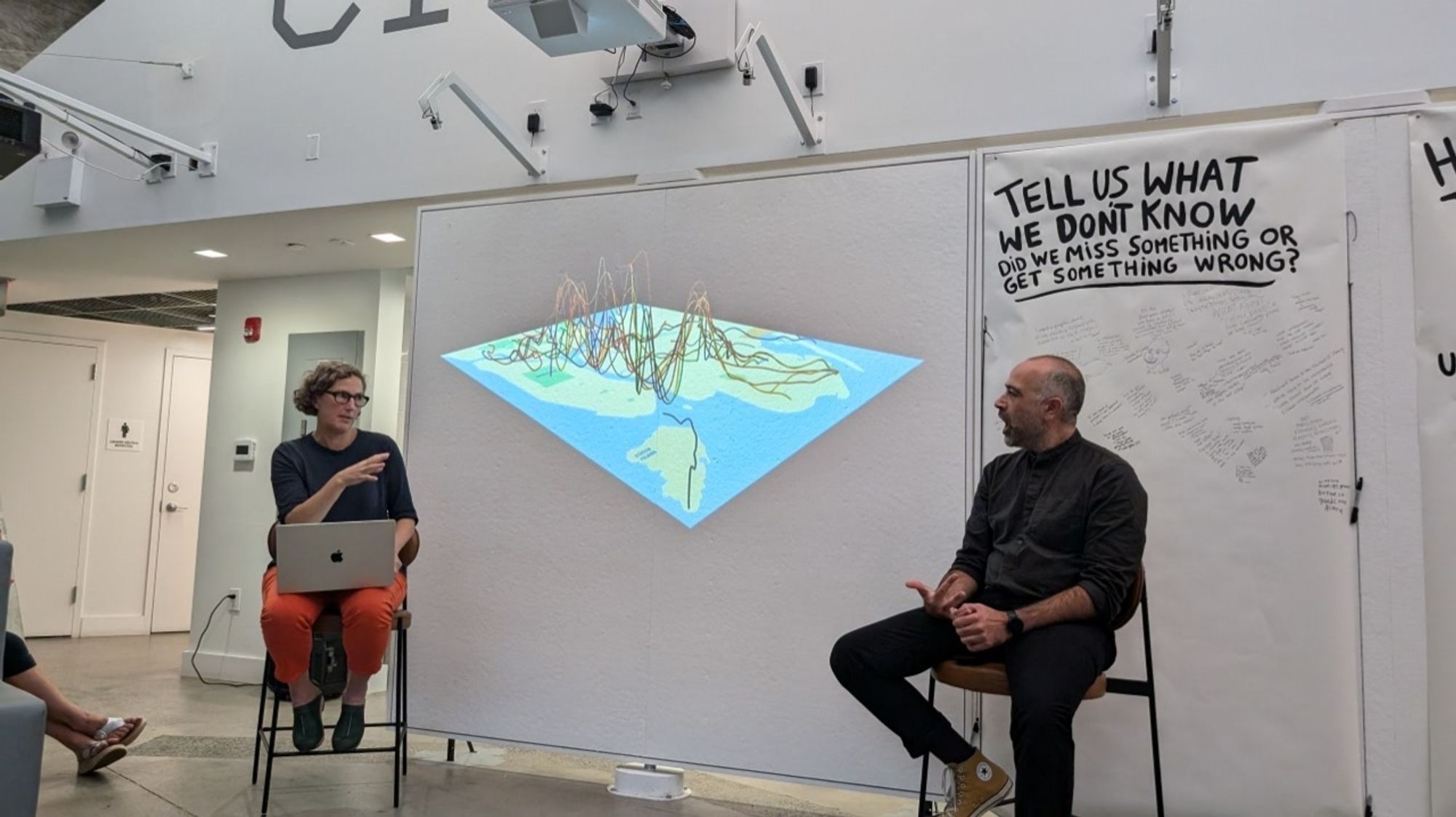 Two people, one woman with a laptop on her lap and a man, sitting on stools on either side of a slide showing a 3D representation of the NYC transit map.