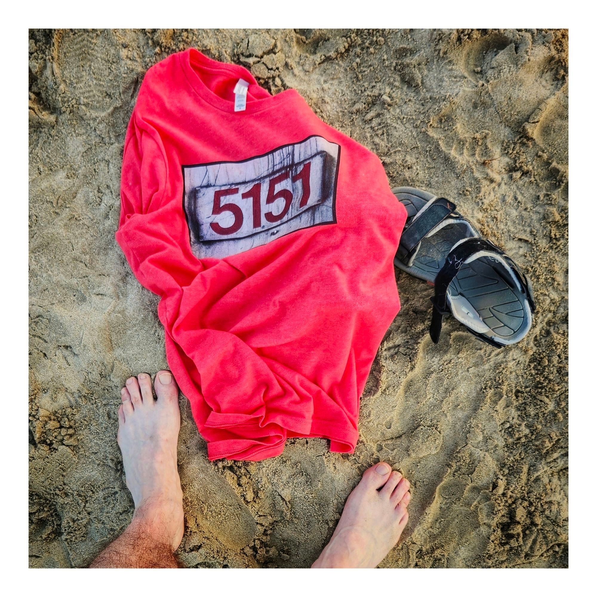 A red t-shirt with 5151 lies on a beach with a sandal and two gnarly feet nearby