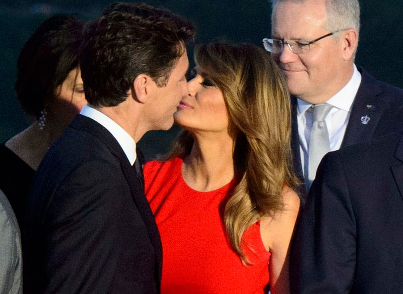 Melania Trump in a bright red dress kissing Justin Trudeau on the lips. A white haired gentleman in a dark suit is standing behind her, smirking because Melania is making Donald look like a fool. 