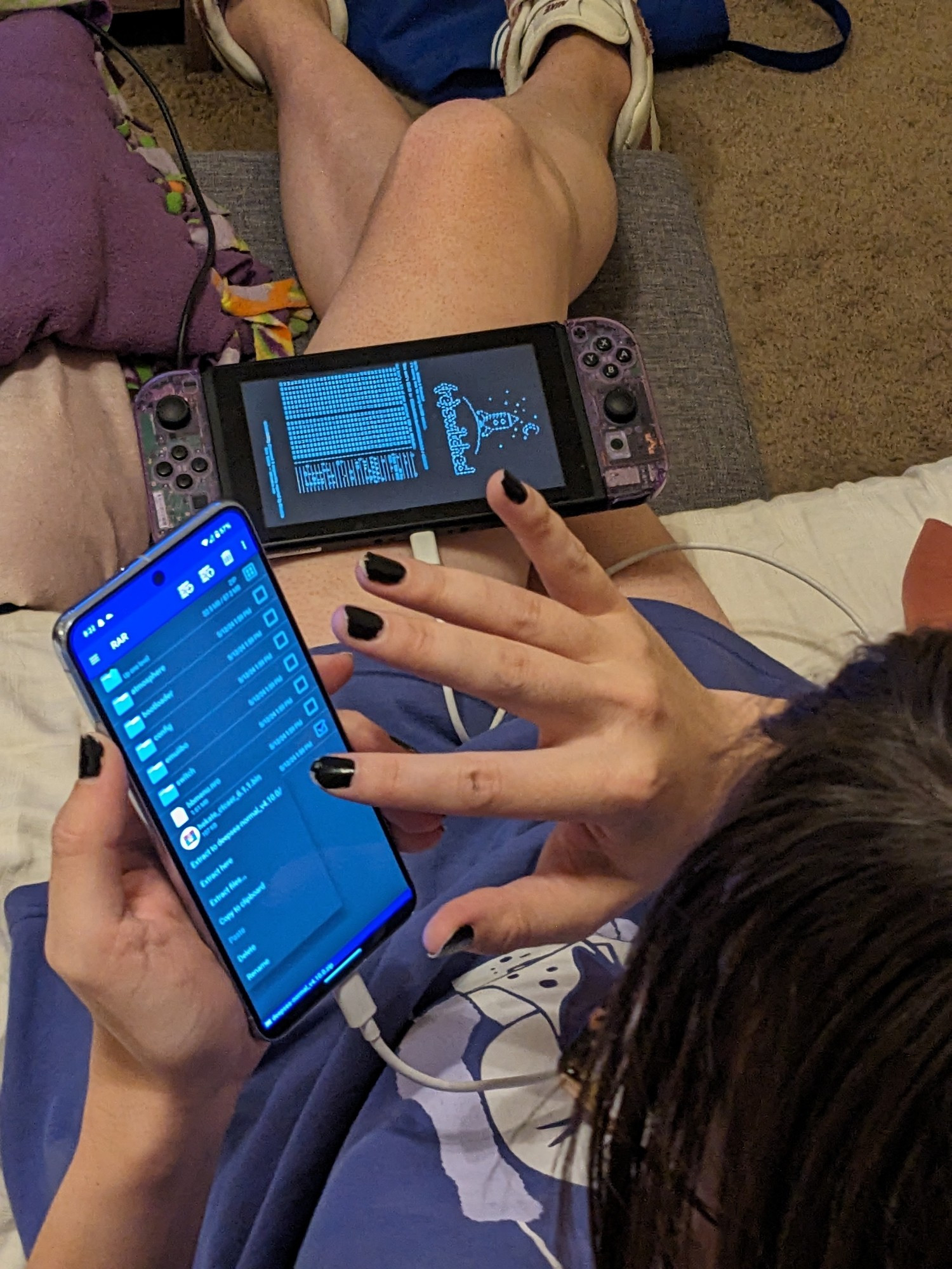 gf is haxxoring a nintendo switch that's resting on her thigh with her cell phone plugged in via usb-c. Her legs are crossed and she has on black nail polish.