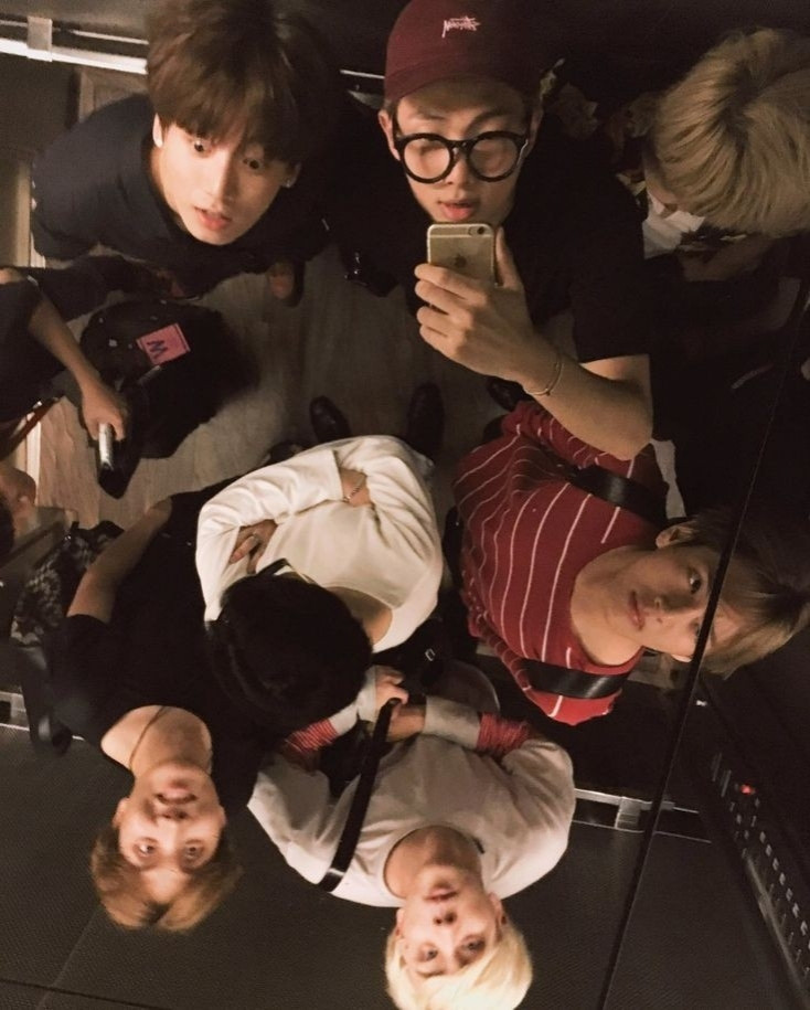 A photo of the group BTS from an overhead angle, everyone is in an elevator and Namjoon is taking a selfie of everyone through the ceiling mirror, some of the boys noticed and some didn't.