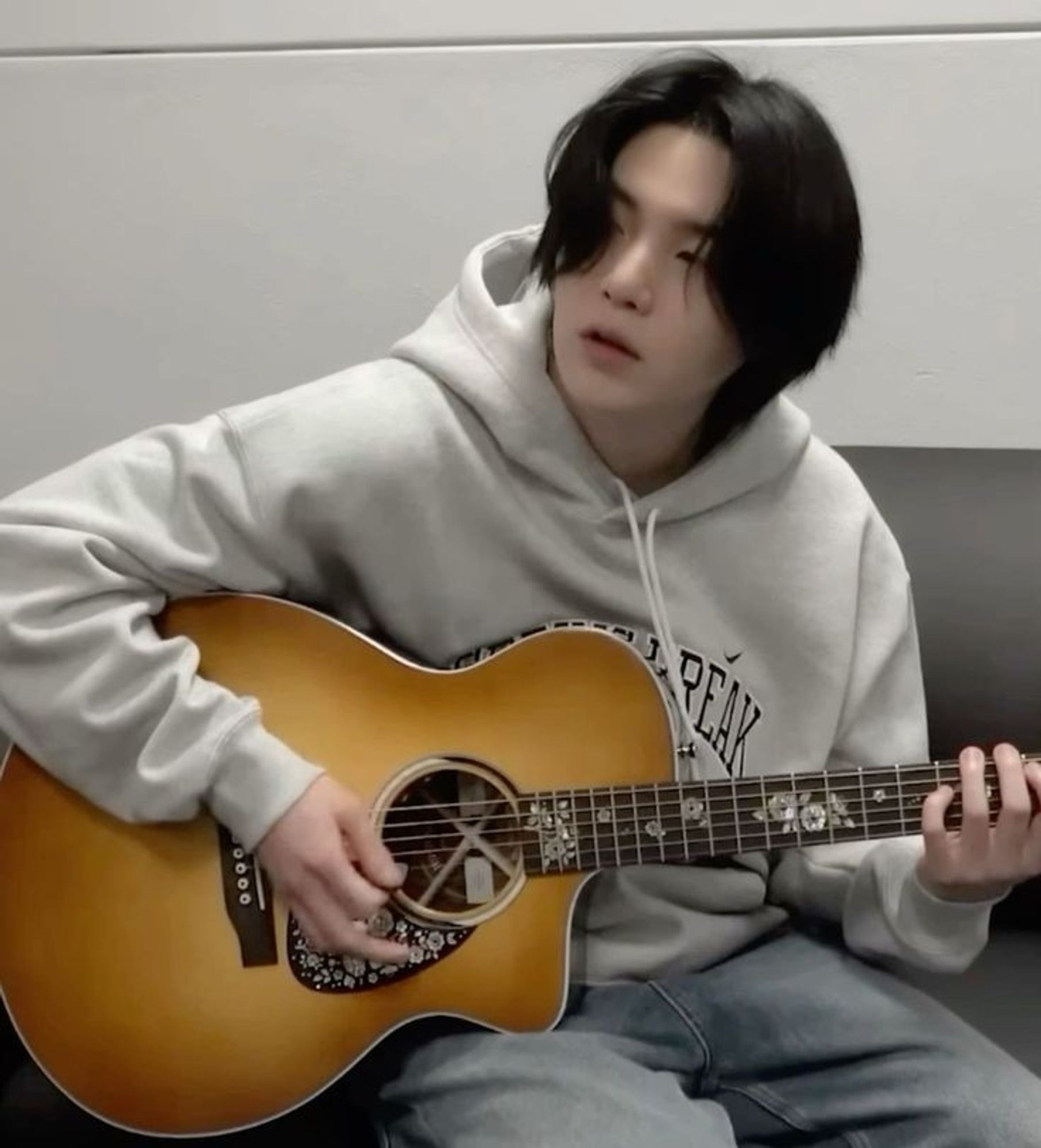 A photo of BTS's Yoongi playing the guitar, he is wearing a light colored hoodie and jeans, his hair is black and the guitar is a light brown shade. The idea of the post is to make a comparison between Snoopy's drawing and Yoongi's photo.