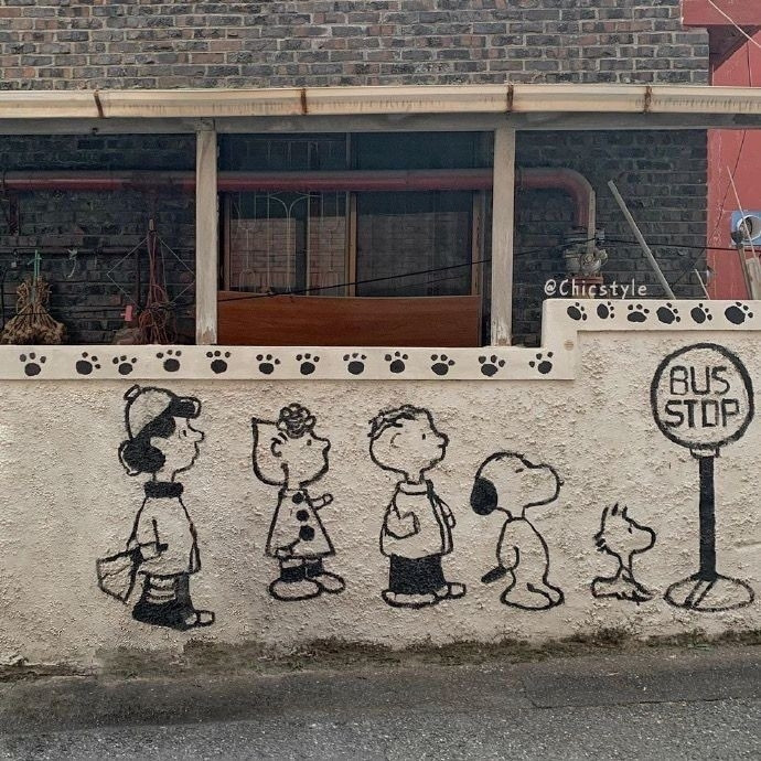 A photo of a wall of a residence where a drawing of Charlie Brown's gang, including Snoopy and Woodstock, is graffitied. They are waiting for a bus side by side and next to them there is a drawn of a bus stop sign.