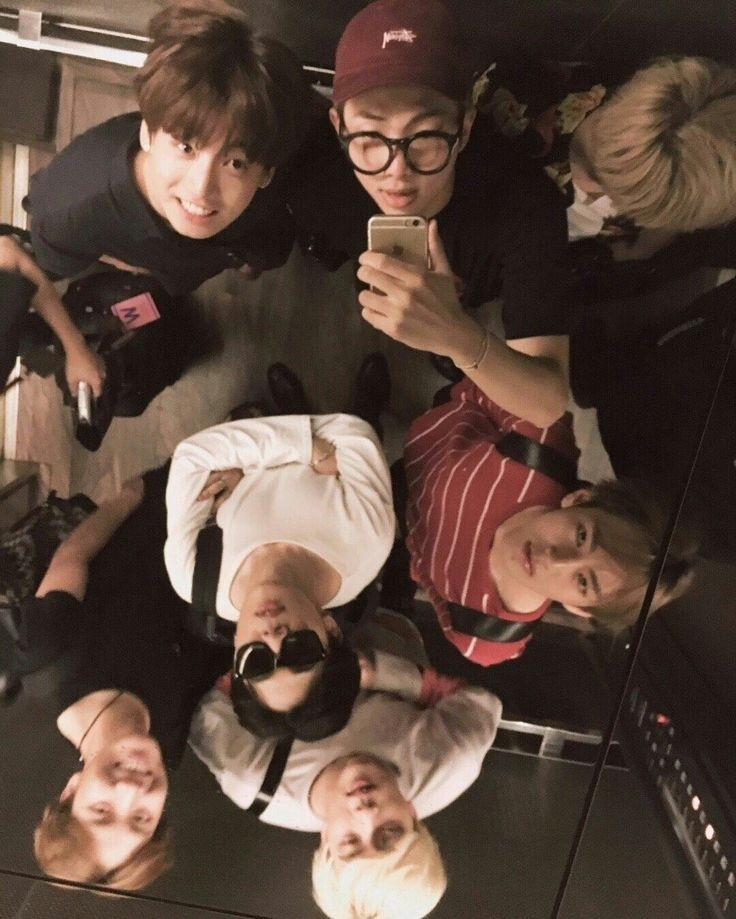 A photo of the group BTS from an overhead angle, everyone is in an elevator and Namjoon is taking a selfie of everyone through the ceiling mirror, some of the boys noticed and some didn't