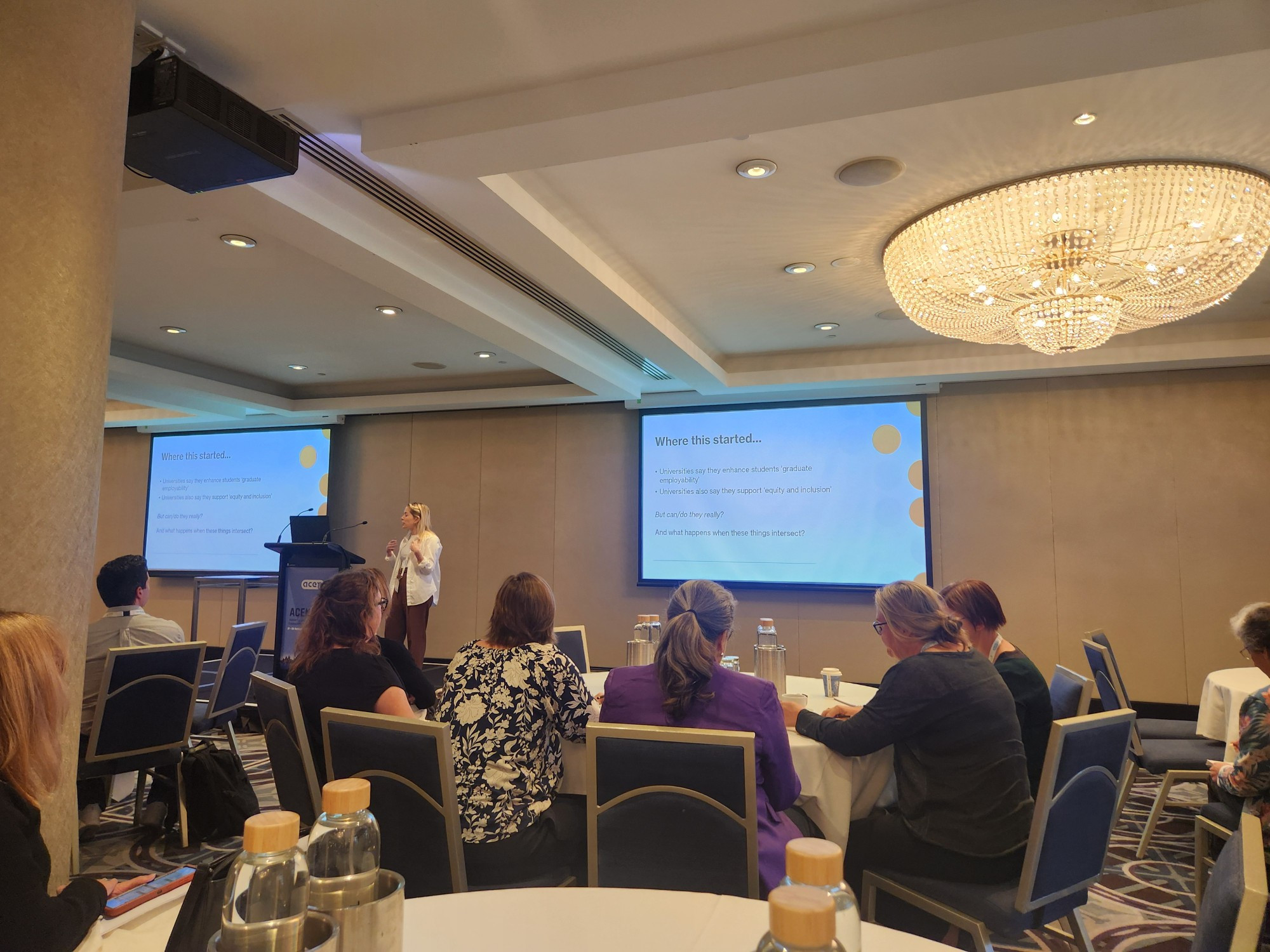 Mollie at the lectern, presenting in a room with huge chandeliers and pillars blocking the view. 