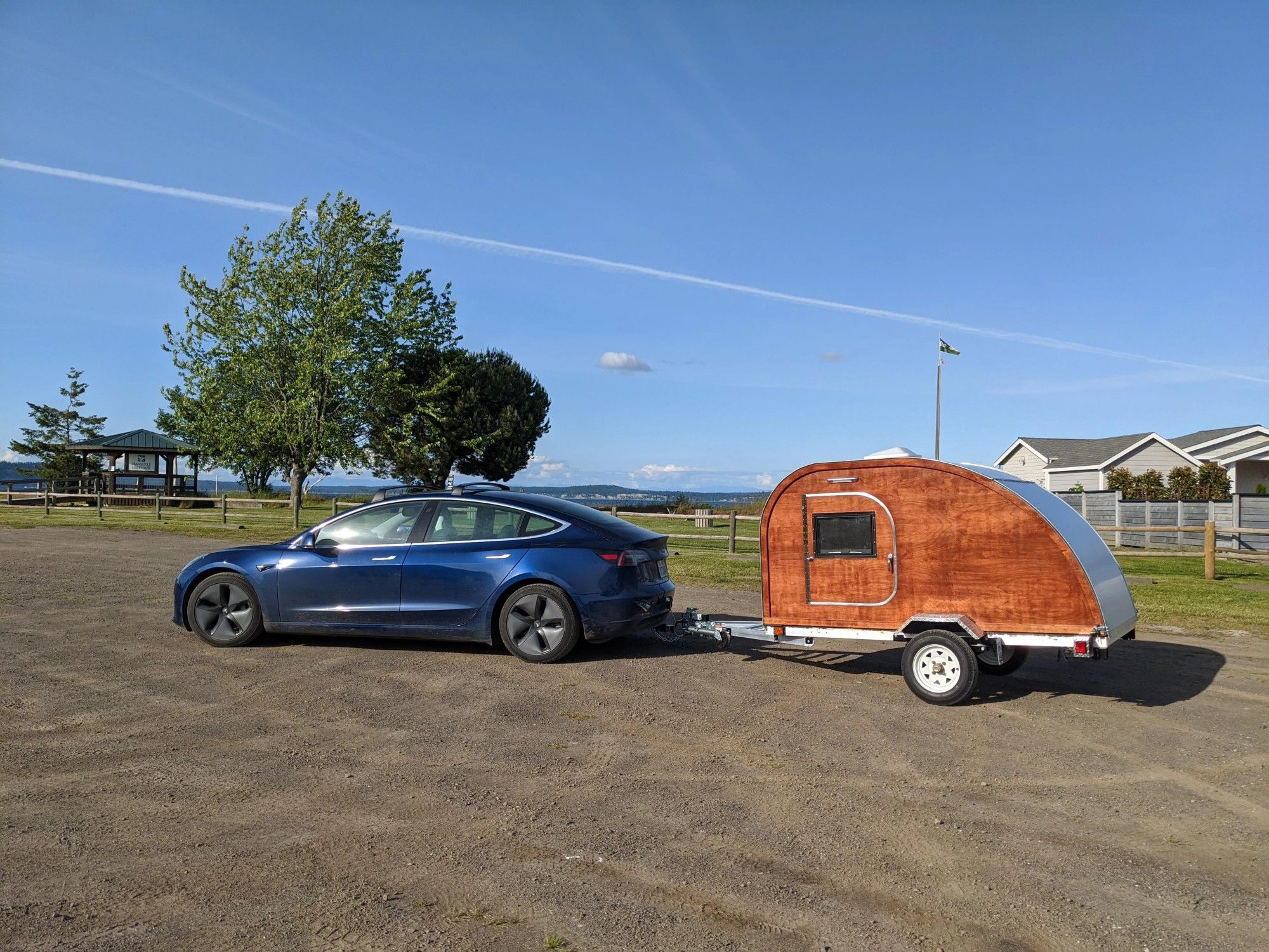 A blue Tesla Model 3 with a wood-paneled teardrop-shaped travel trailer on a towing hitch. It's in the parking lot of what appears to be a park in coastal Washington State.