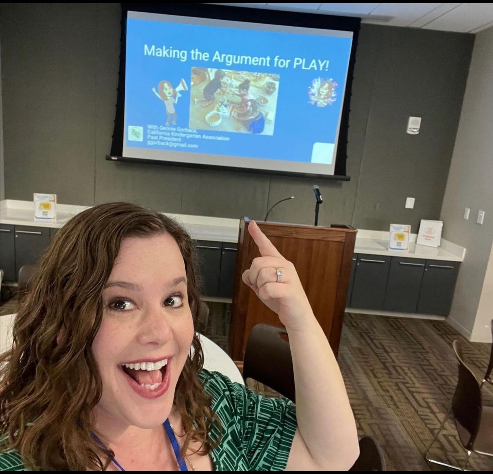 Photo of smiling Gennie wearing a book themed dress and pointing to a screen featuring the opening slide of one of her professional development sessions, entitled “Making the Argument for PLAY!”