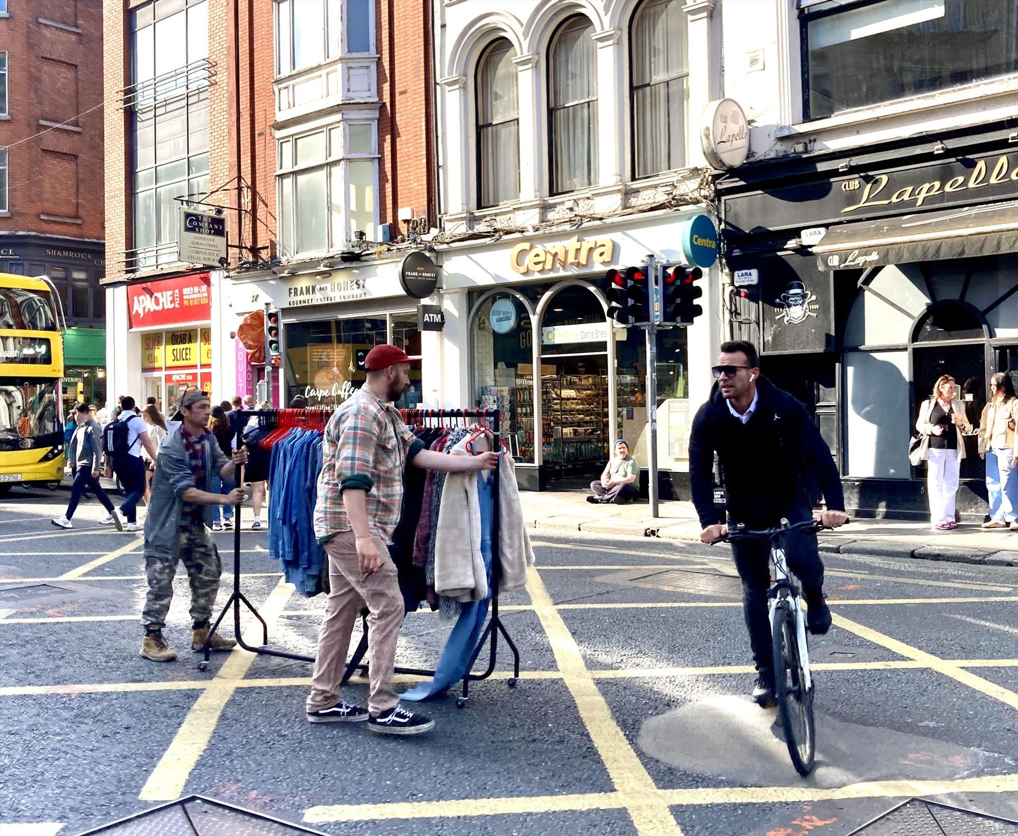 Two men push a rack of hanging clothes across Dame Street while another man on a bicycle dodges them. In the background, a river of pedestrians crosses at a light, while a bus waits.