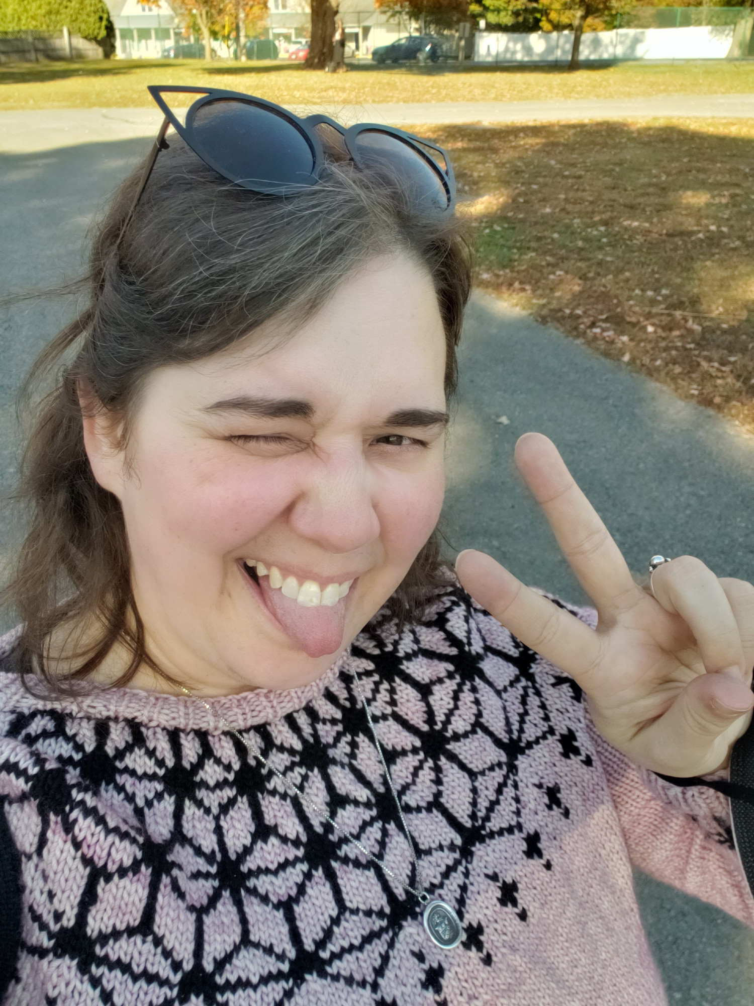 a woman outside on a sunny day, wearing a handknit pink sweater with a black pattern, making a peace sign and sticking out her tongue while squinting at the sun