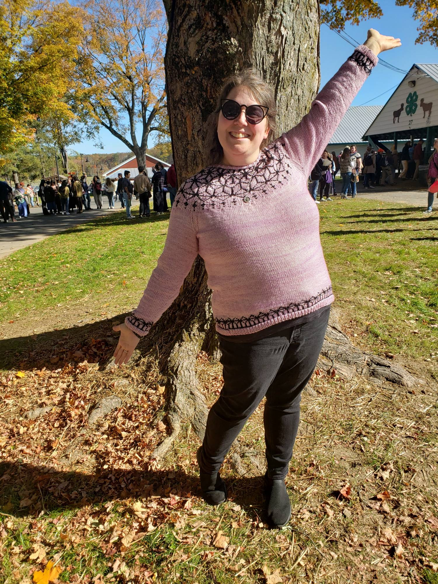 a woman outside on a sunny day, wearing a handknit pink sweater with a black pattern, her arms outstretched as she smiles