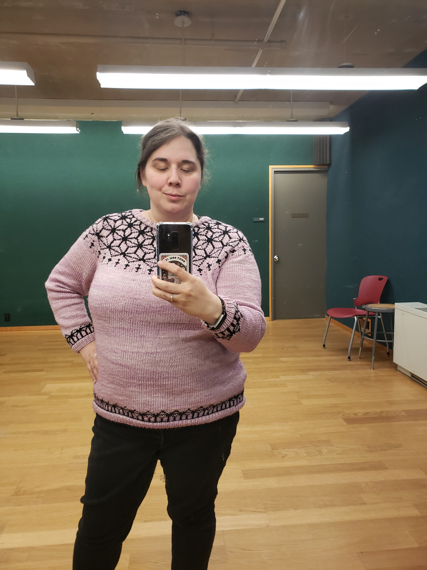 a woman standing in a room with green walls, wearing a handknit pink sweater with a black pattern, taking a mirror selfie 