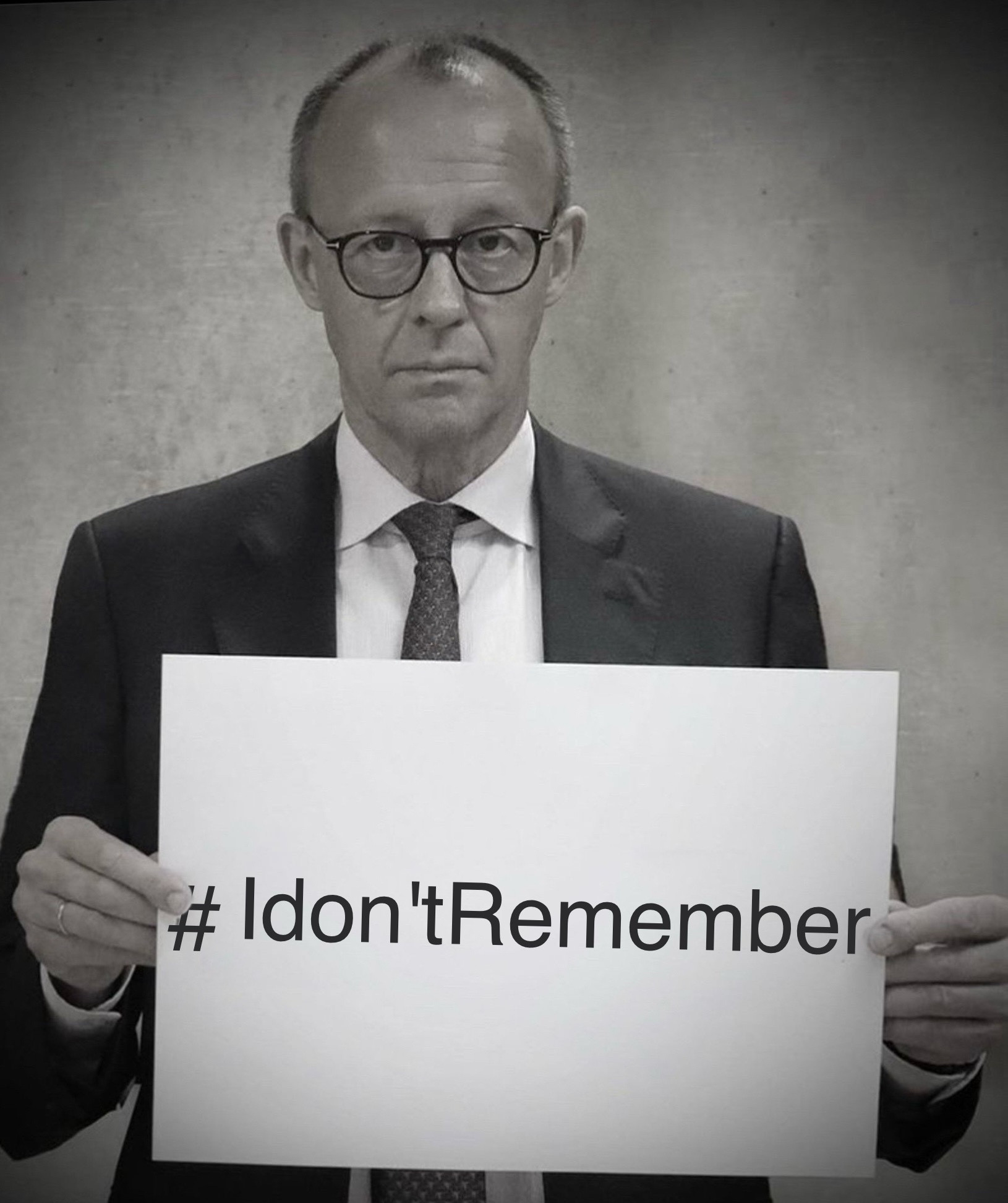 Ein s/w Bild von Friedrich Merz.
Er hält mit bemüht betroffenem Gesichtsausdruck ein Schild hoch.
Im Original, auf seinen Social Media Accounts verbreitet, steht darauf #WeRemember.

Der Schriftzug wurde auf #Idon'tRemember geändert.