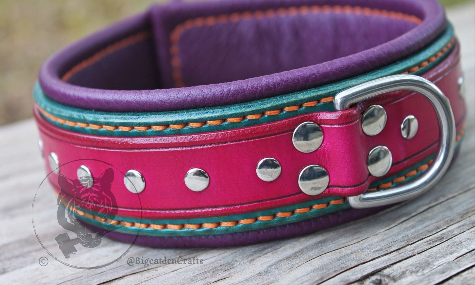 Close-up of a multi-layered leather collar and short leash sit on a wooden board. The collar has a fuchsia top strap with silver-colored rivets studding its surface, turquoise/teal back strap, plum purple interior lining, orange stitching, and stainless steel hardware.