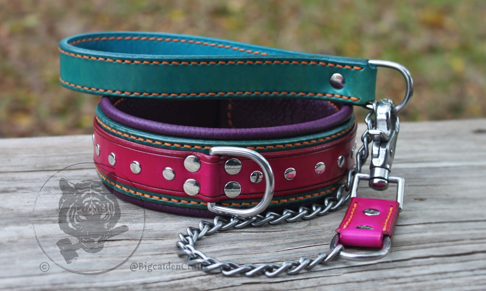 A multi-layered leather collar and short leash sit on a wooden board. The collar has a fuchsia top strap with silver-colored rivets studding its surface, turquoise/teal back strap, plum purple interior lining, orange stitching, and stainless steel hardware. A matching short leash has a turquoise/teal leather handle, fuchsia leather grip behind the clasp, orange thread, steel chain, and stainless steel d rings and clasp.