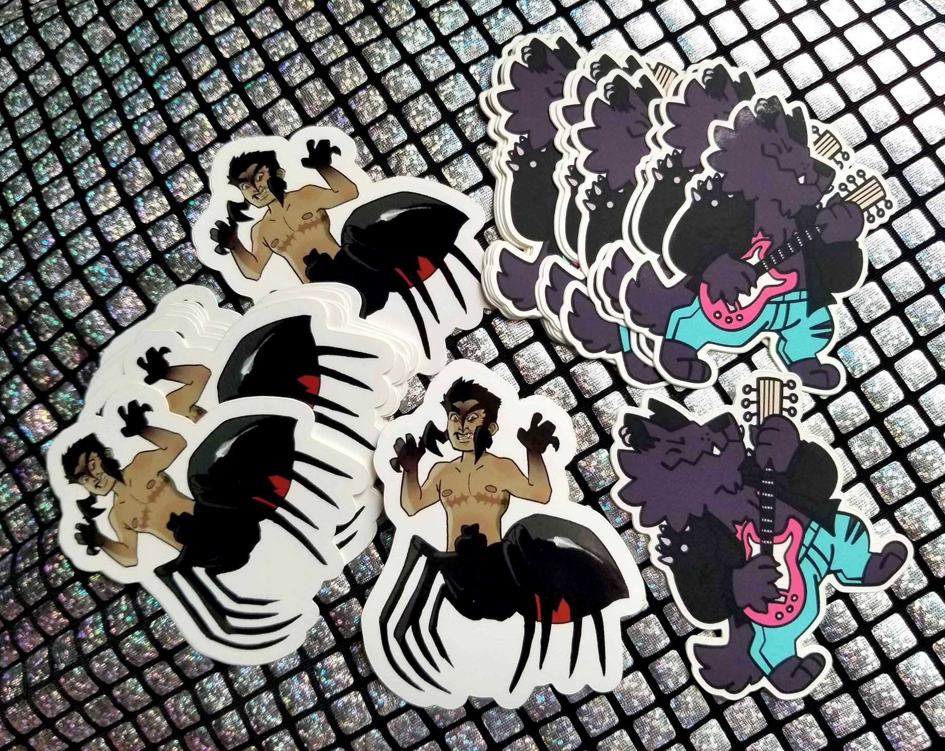 Photo of two small piles of die-cut stickers on a shiny backdrop. On the left is a black widower--a character that resembles a drider with a humanoid top and spider bottom, but they are transmasculine with a black widow body. On the right is a dark furred werewolf in a leather jacket rocking out on an electric guitar.