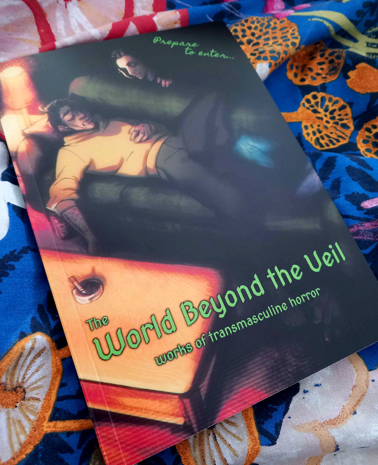 Photo of The World Beyond the Veil zine of transmasculine horror, showing the cover with a transmasc individual laying on a couch while a shadowy figure leans over them.