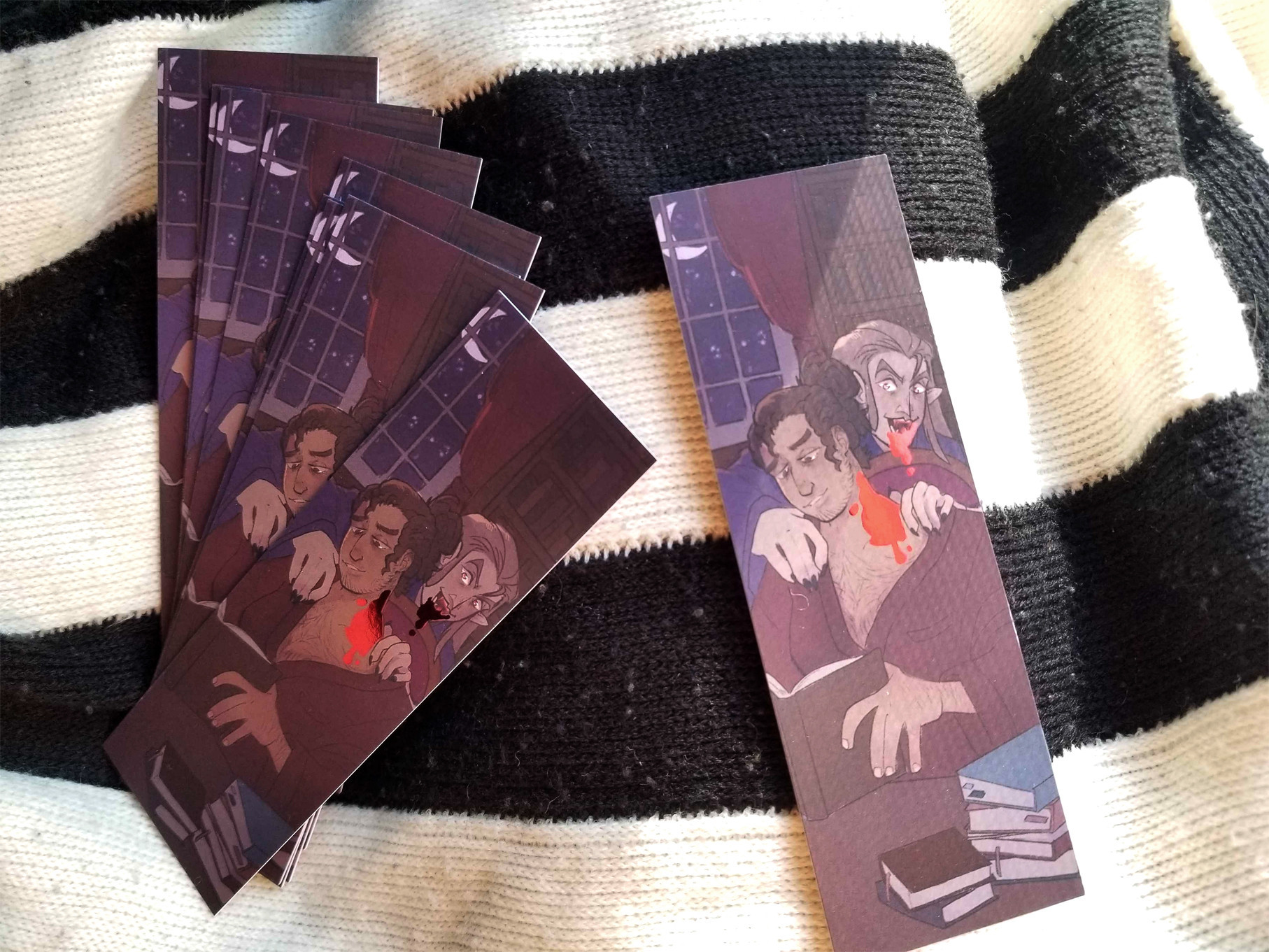 Photo of some bookmarks on a striped backdrop. The bookmarks feature two masculine figures, a vampire feeding on a human who's reading from a stack of books. The blood on the human's neck is a reflective foiled spot, shining in the sunlight.