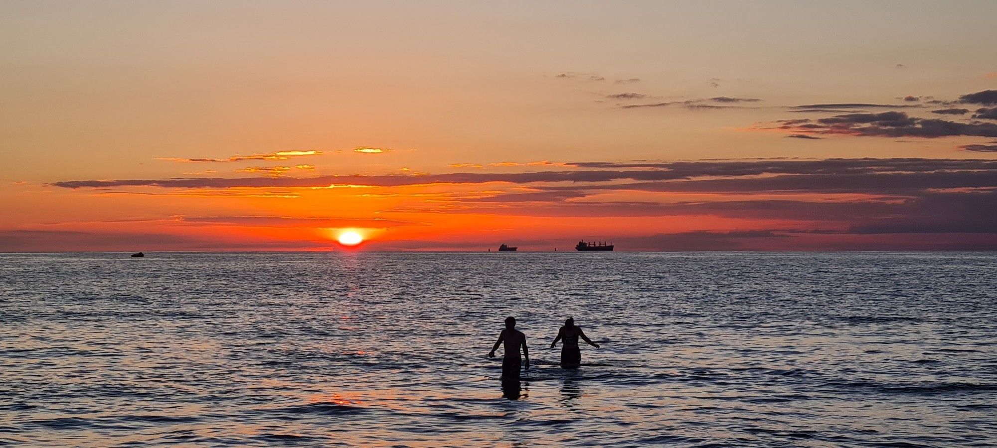 Sonnenuntergang mit Abendrot am Ostseestrand, im Wasser die Silhouette von zwei Personen.