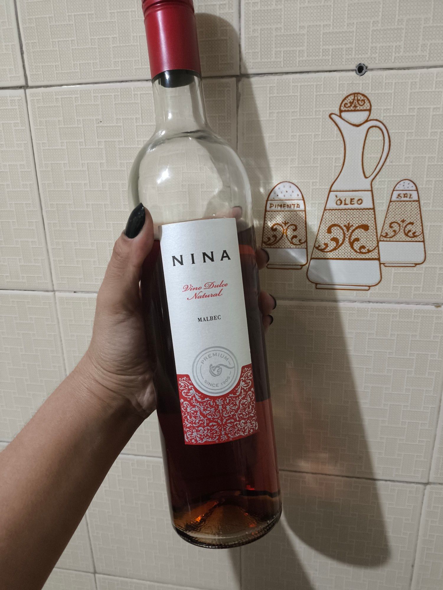 Garrafa de vinho  Nina argentino Malbec Doce Natural, de cor rosada, rótulo branco e vermelho. Ao fundo, parede azulejos bege decorado com motivos de culinária.