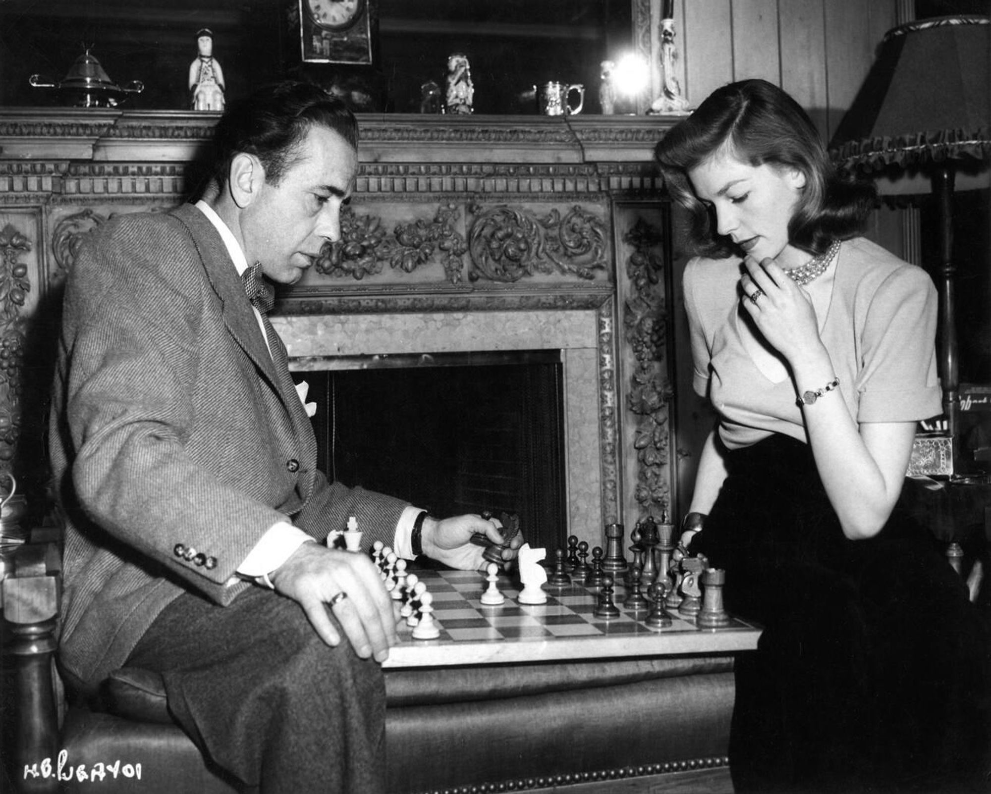 Actors Humphrey Bogart and Lauren Bacall — Bogart dressed in a dark-coloured suit jacket, white shirt, dark-coloured trousers and bow tie, Bacall in a two-toned gown with light-coloured top and black skirt — captured sitting across from each other in profile, a chessboard between them, both looking intently at the pieces as Bogart lifts a knight and a pawn from Bacall's material, in a medium-length black-and-white shot taken in their home at Benedict Canyon in Los Angeles