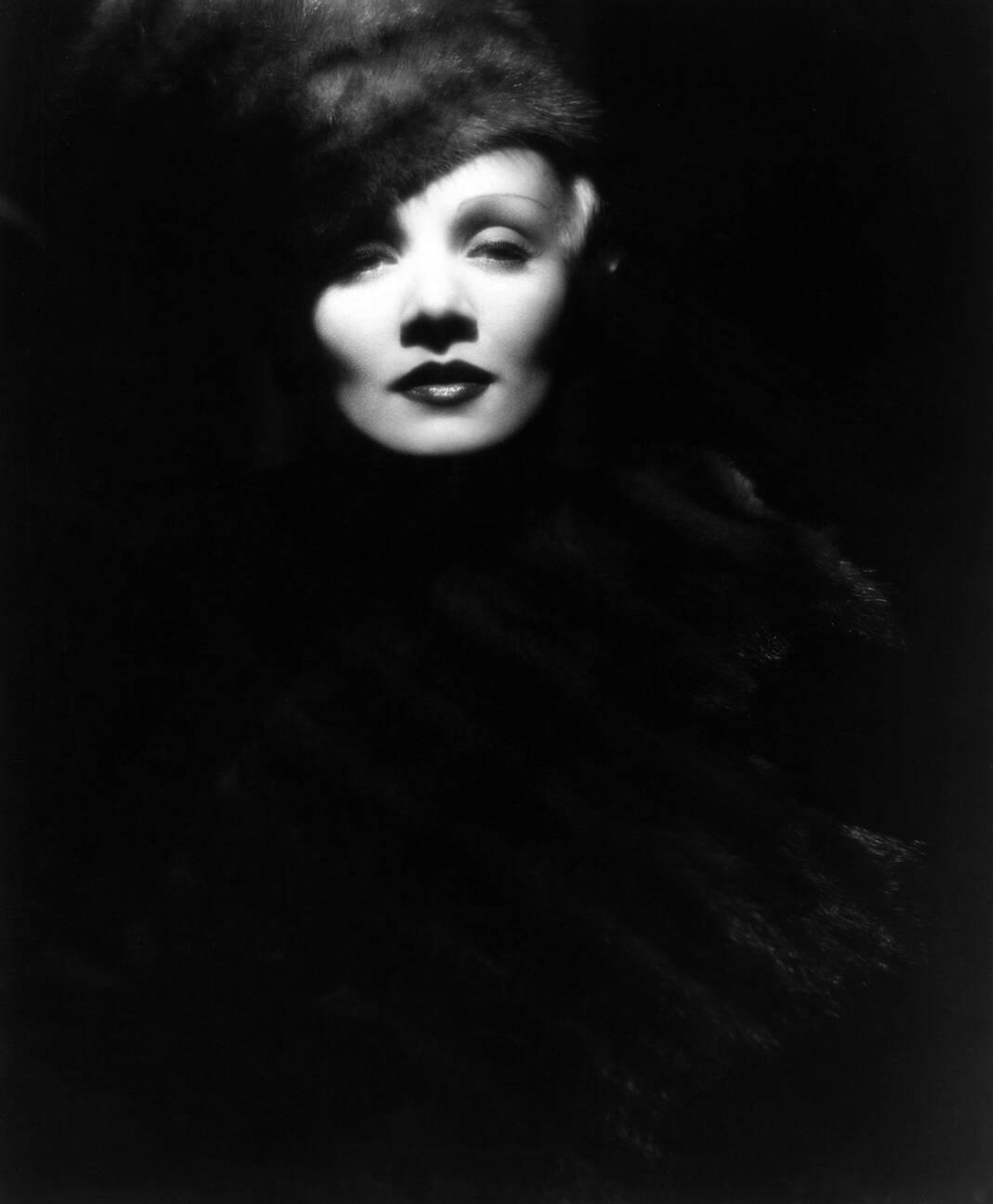 Actress and singer Marlene Dietrich — dressed in a black high-necked gown, dark-coloured fur hat, wrapped in an extravagant dark-coloured fur stole — captured facing the camera, her visage cloaked mostly in shadow in an atmospheric medium-length black-and-white shot for the film The Scarlet Empress (1934)