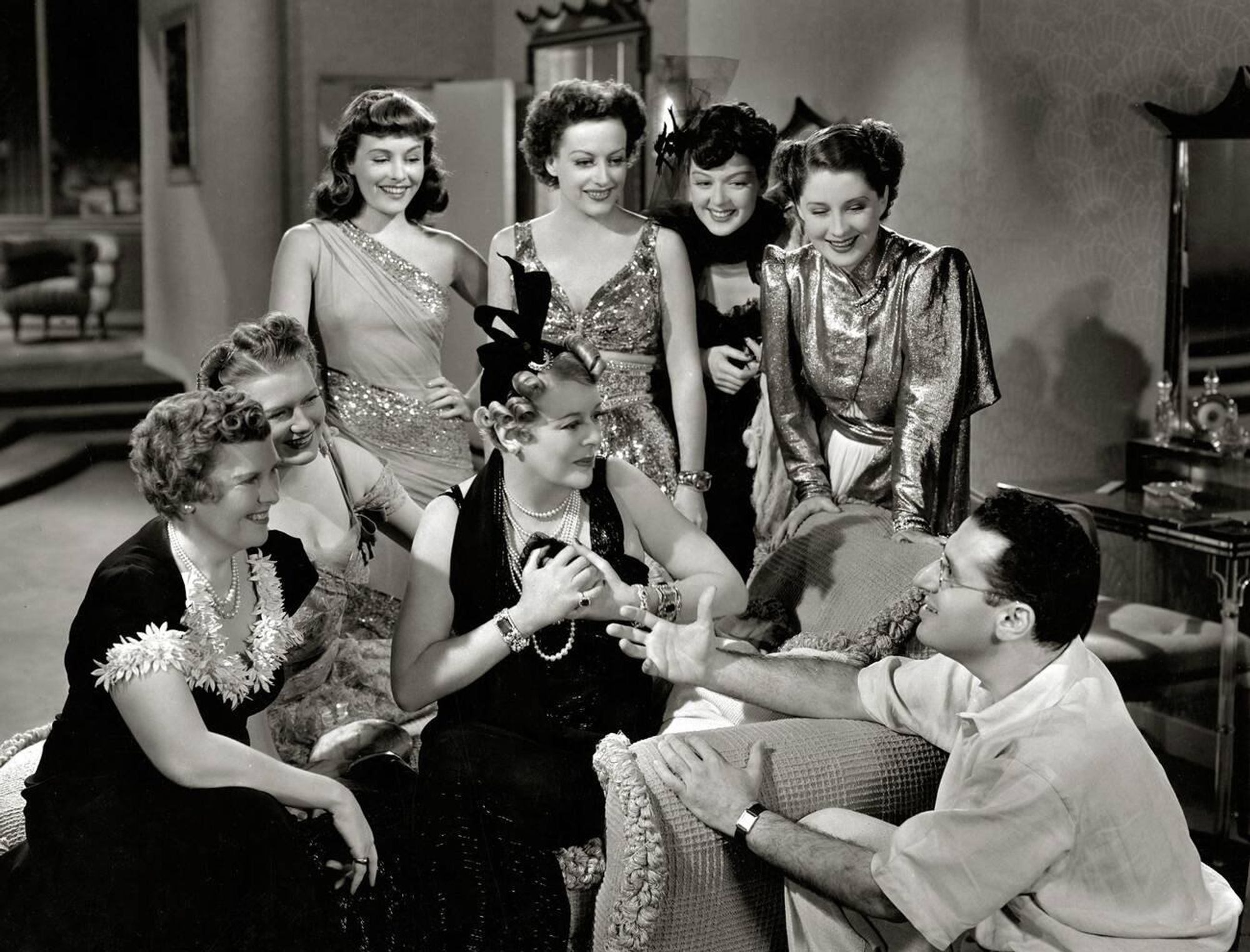 Actresses Paulette Goddard, Joan Crawford, Rosalind Russell, Norma Shearer, Florence Nash, Phyllis Povah, Mary Boland and director George Cukor — the women all dressed in elegant gowns designed by Adrian, Cukor in a light-coloured casual shirt and trousers — captured together, Nash, Povah and Boland seated as Goddard, Crawford, Russell and Shearer stand behind them, all looking down at Cukor, who crouches on the floor and looks toward Nash, all smiling, in a medium-length black-and-white shot taken during a break from filming on the set of The Women (1939)