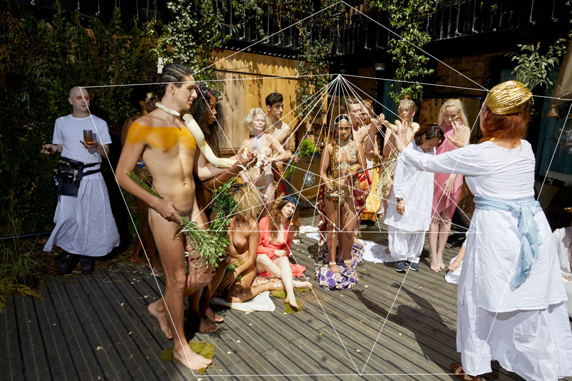 A moment from Mandrake—the Apple of Love, a performance by Iris Brosch, written by Edward Peacock : a crowd mill about in a hotel's open atrium under leaf-dappled sunlight, a naked man, smeared with orange, with a yellow python about his shoulders, a woman crowned in gold in a white dress, a pale blue sash about her waist, other figures in various states of dress or undress, and a mostly symmetrical shape of thin white lines radiating from this point of the scene, or that, suggesting interlocking connections of an arcane nature between and among them all.