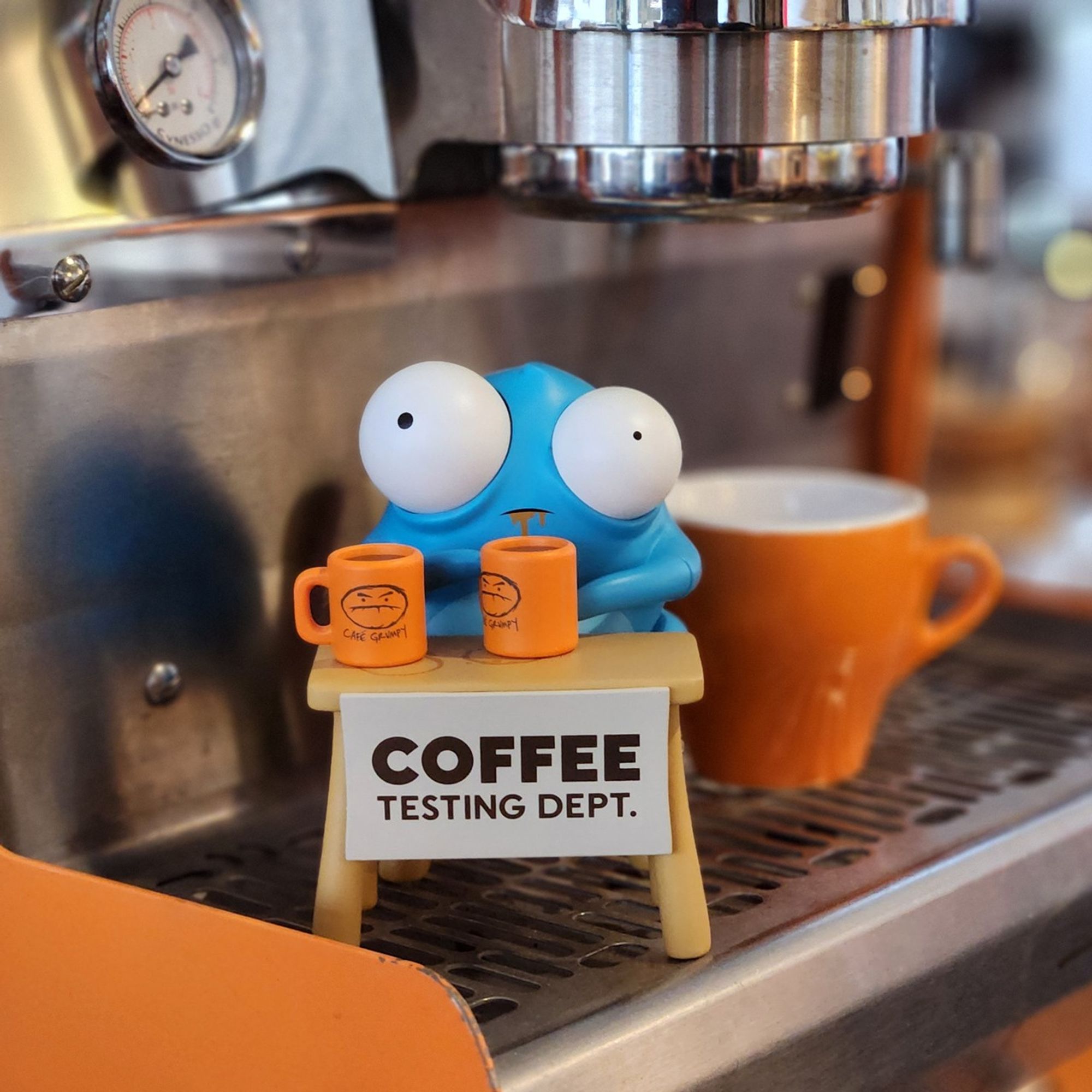 Picture of blue bug-eyed creature sitting on an espresso machine with mugs of coffee, toy by Andrew Bell