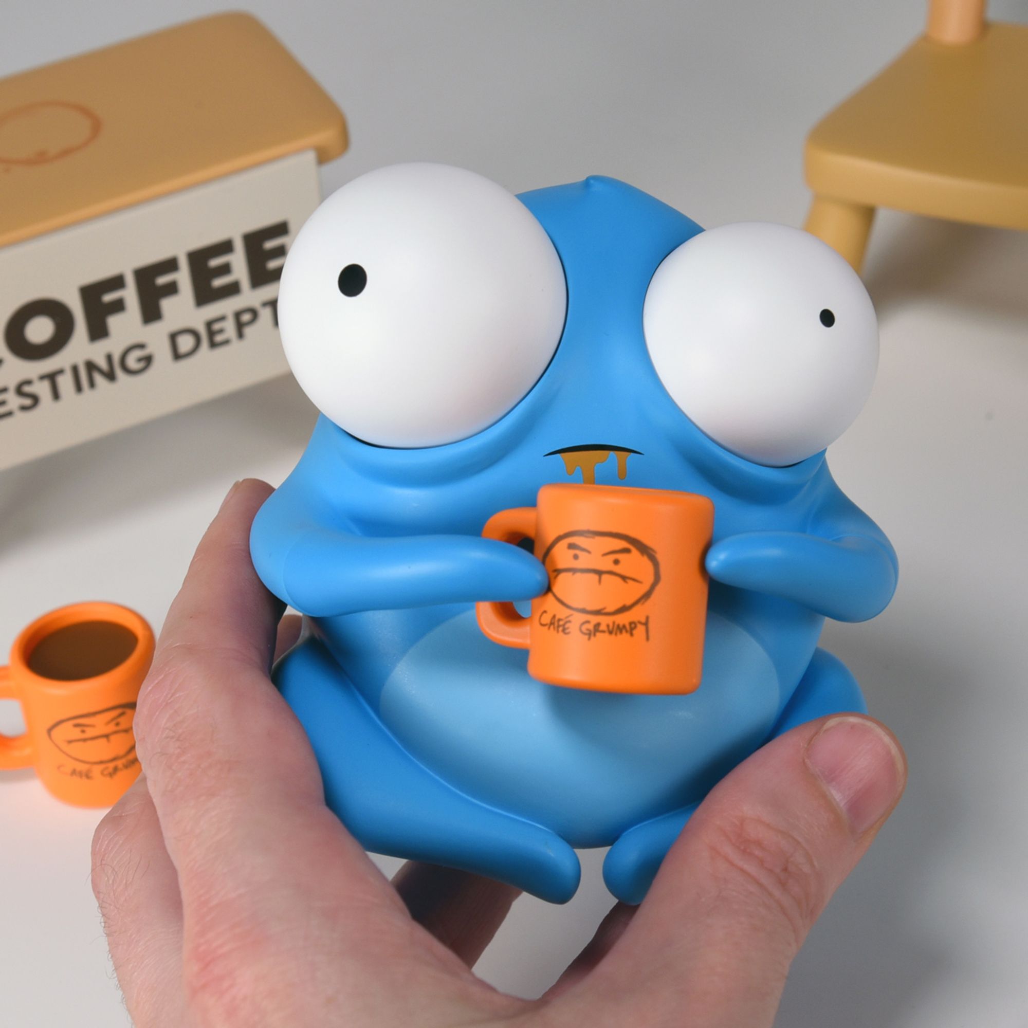 Picture of blue bug-eyed creature being held for scale, it's holding a small mug of coffee with the Cafe Grumpy Logo on it