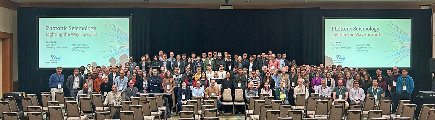 SSA 2024 Photonic Seismology attendees standing in a group on stage at the meeting in Vancouver Westin ballroom with Photonic Seismology slides on either sides of the group