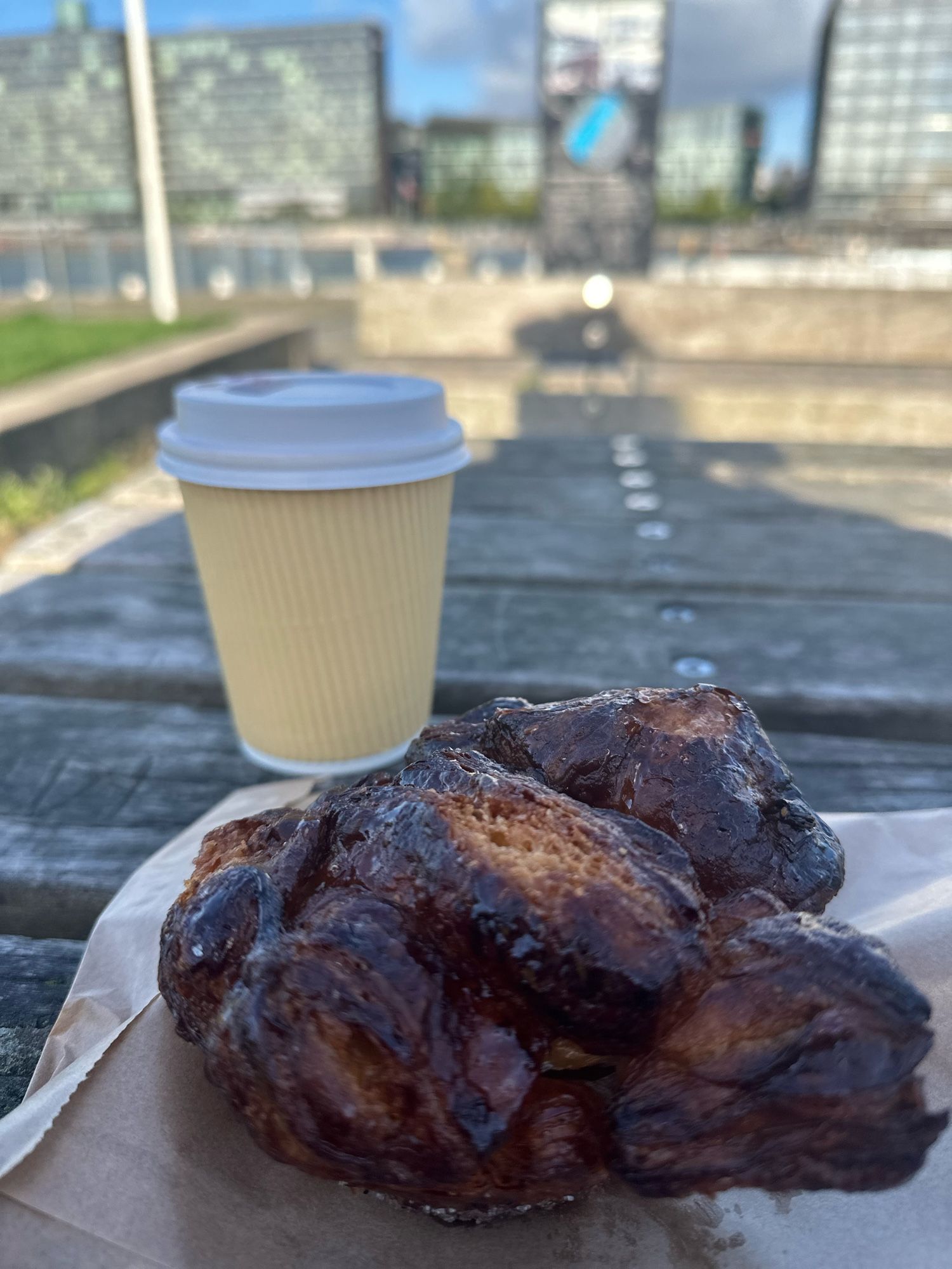 A flaky, dark, cardamom croissant rests on a picnic table with a yellow espresso cup in the background