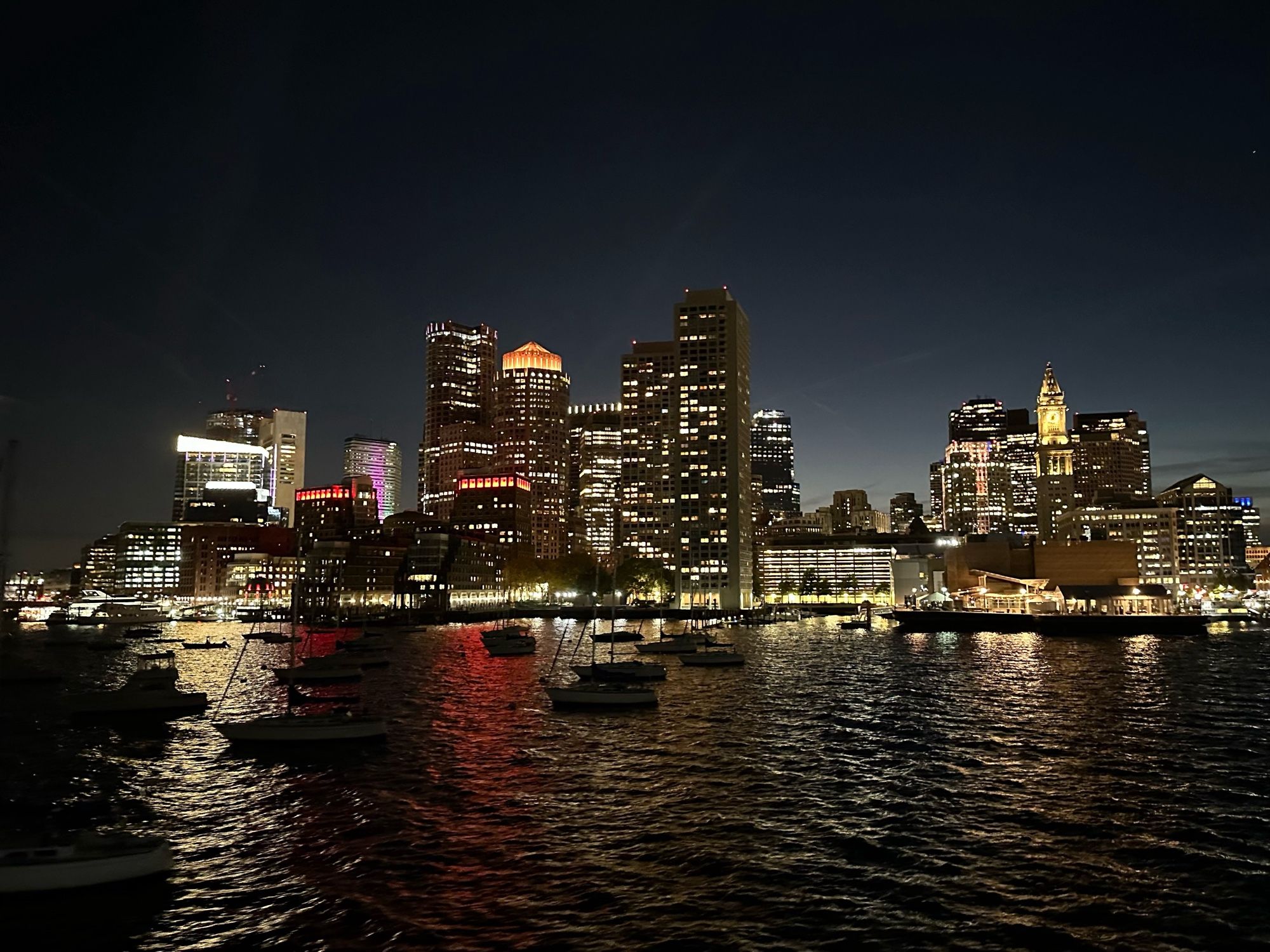 Photograph of the city skyline in Boston, Massachusetts after the sun has set, such that the building lights stand out against the dark evening sky. The view is from a ship in the harbor.