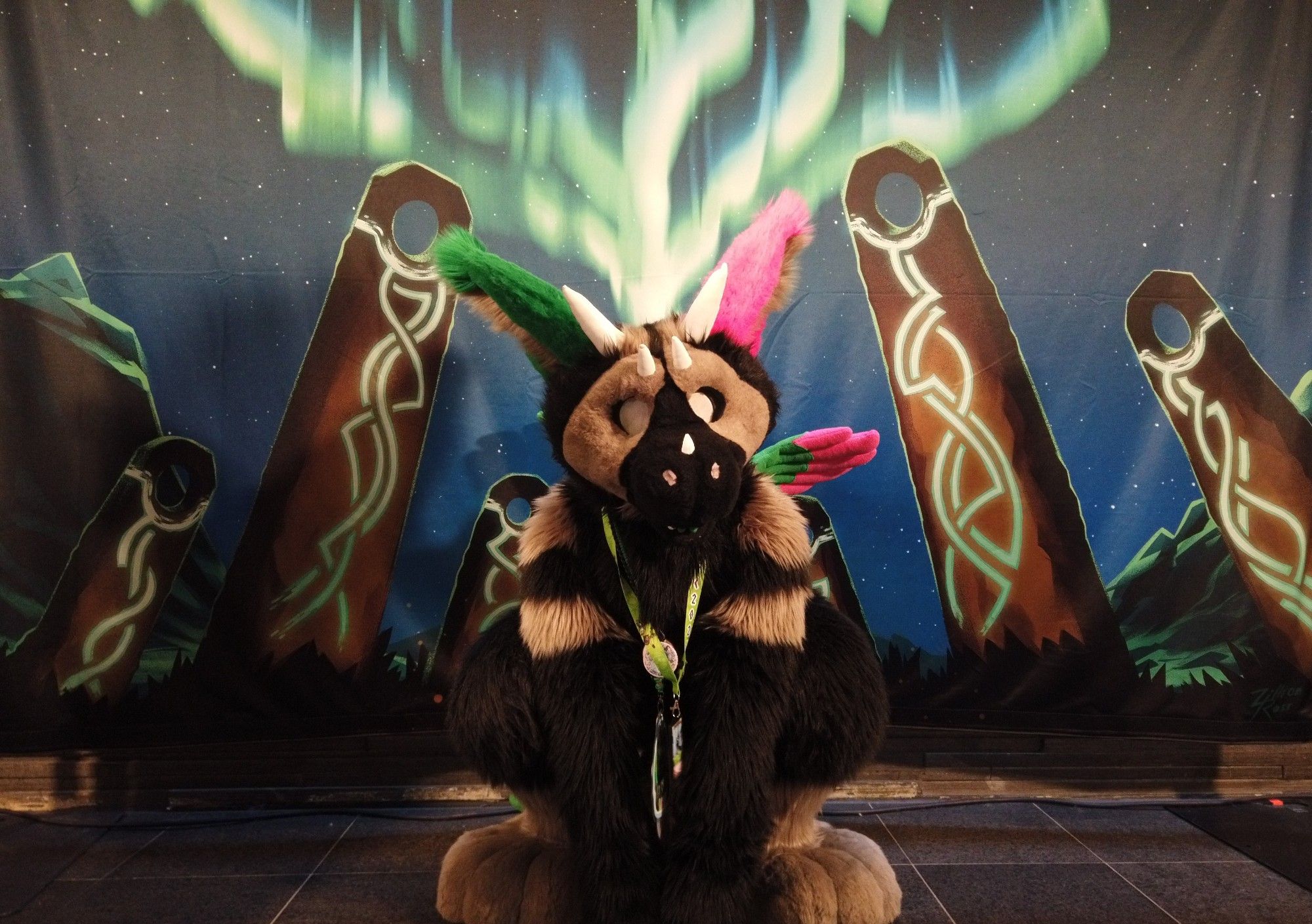 A Dutch Angel Dragon fursuiter squatting in front of a fabric backdrop featuring standing stones, mountains and aurora borealis.