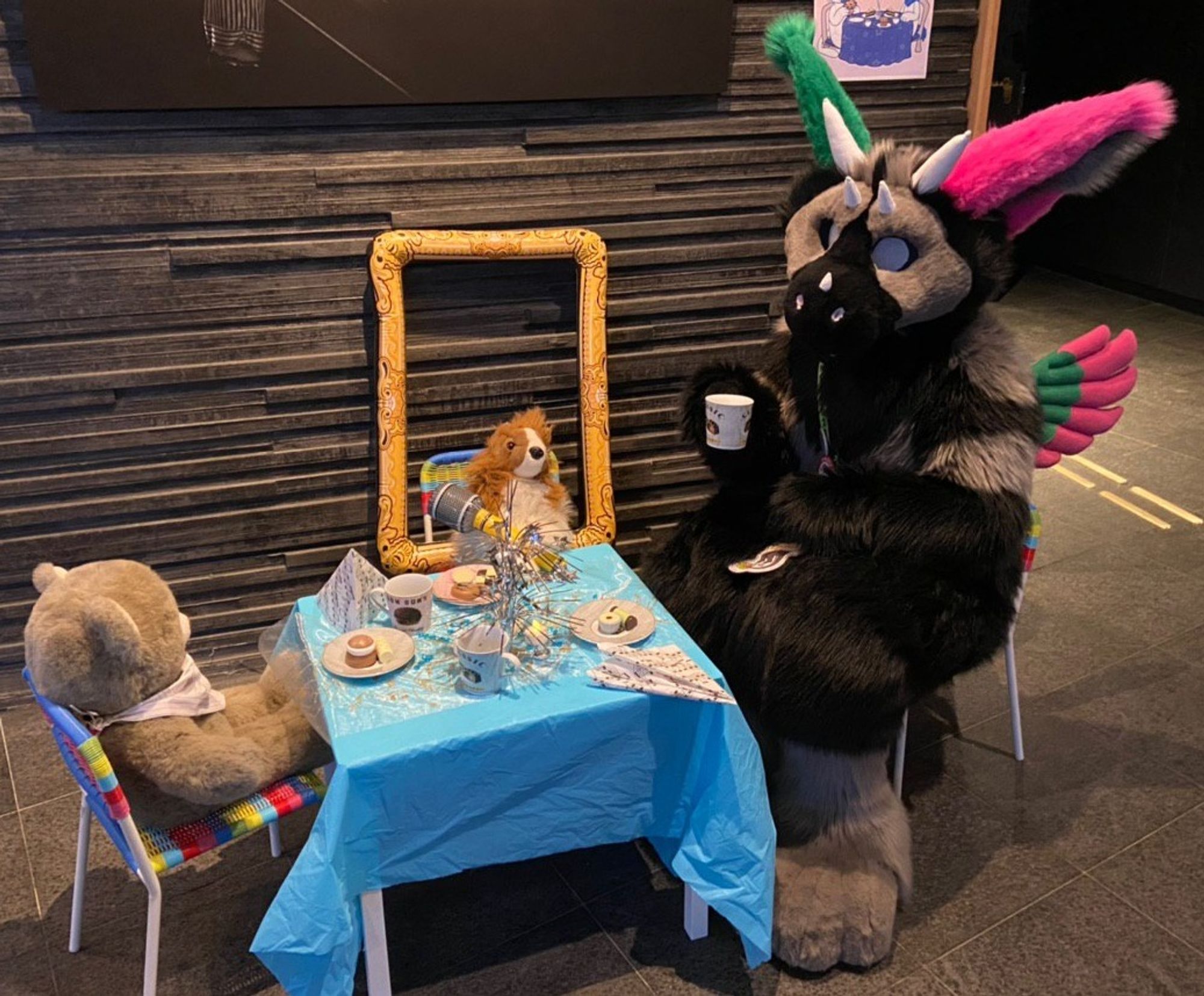 A very small table and chairs laid out with teacups and cakes for afternoon tea. Binky (Dutch Angel Dragon fursuit) is sitting on one tiny chair holding a small teacup, there are soft toys seated on the other chairs.