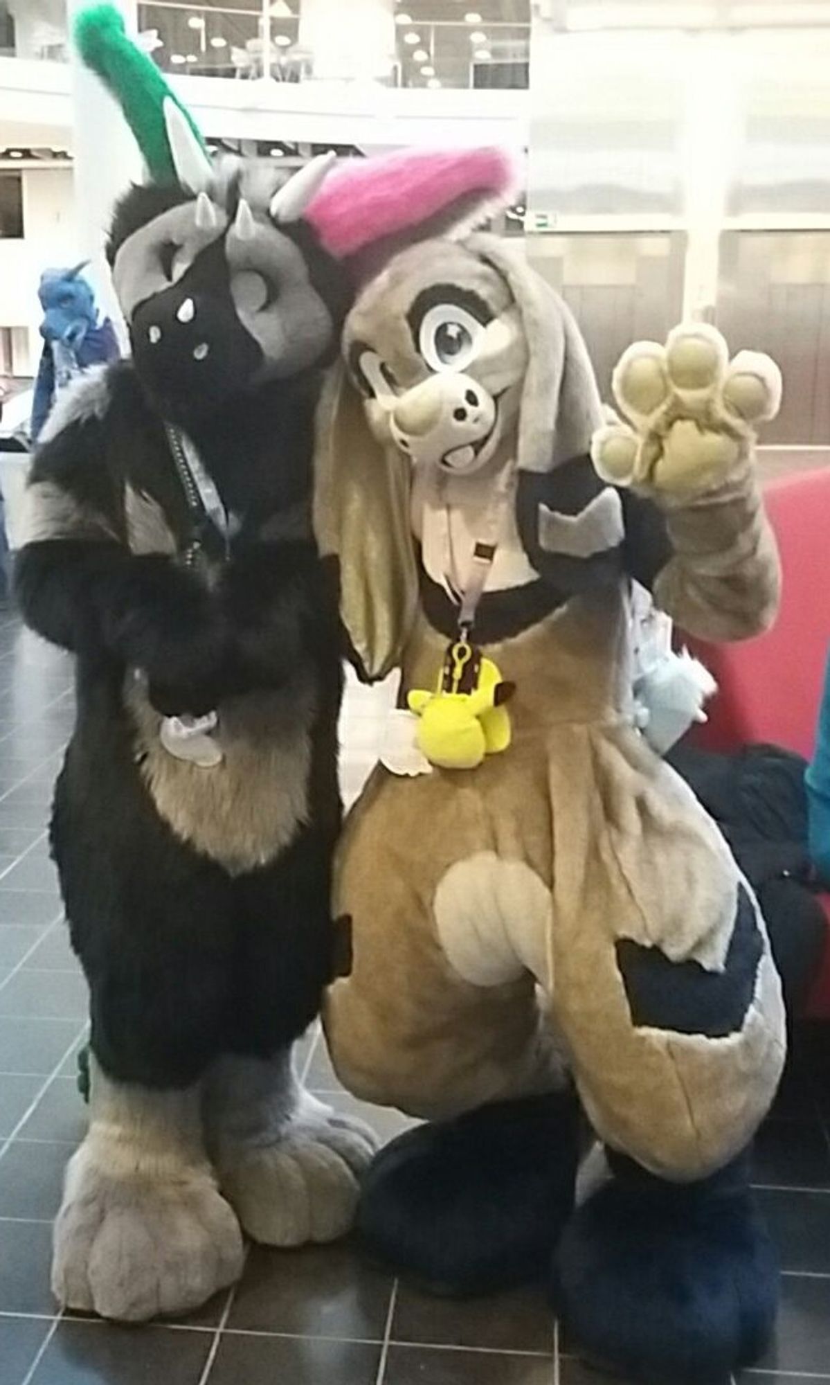 2 fursuiters posing for a photo in the lobby of a hotel. A black and grey Dutch Angel Dragon and a tan and brown lop rabbit who is waving at the camera.