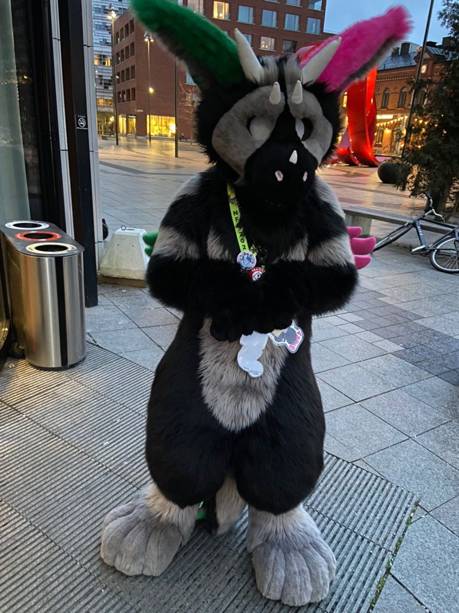 A Dutch Angel Dragon fursuit in black and grey with green and pink details poses outside a hotel. There are illuminated city buildings and a metal modern art sculpture in the background.