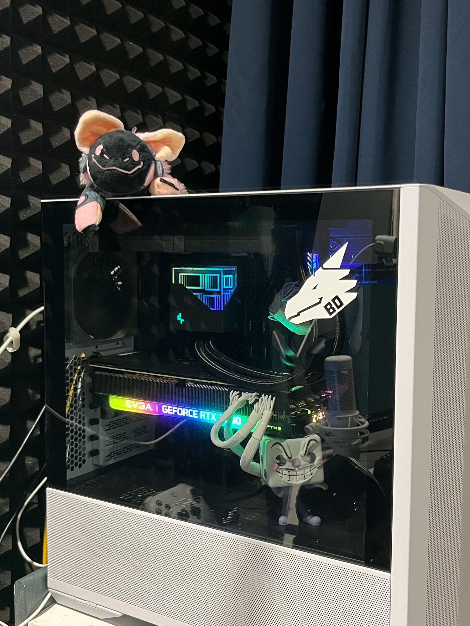 A white and black pc from the side, with rainbow internals. A King Dice pop figure supports the sagging 3080, a bad dragon decal is on the glass of the case, a microphone and controller is reflected off the glass…oh and a pink protogen stuffed toy rests on top of the pc!