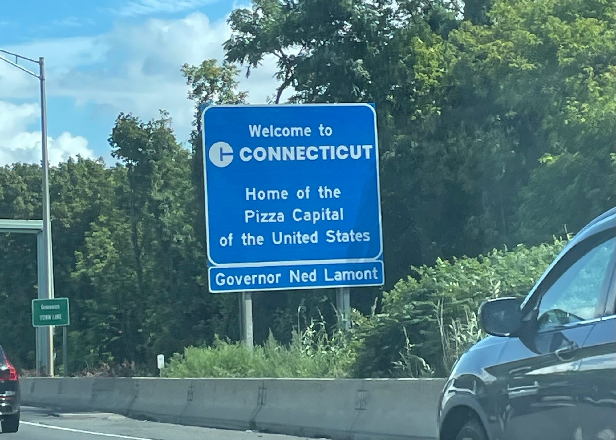 A photograph of a blue sign on a highway that says “welcome to Connecticut, home of the pizza capital of the United States”
