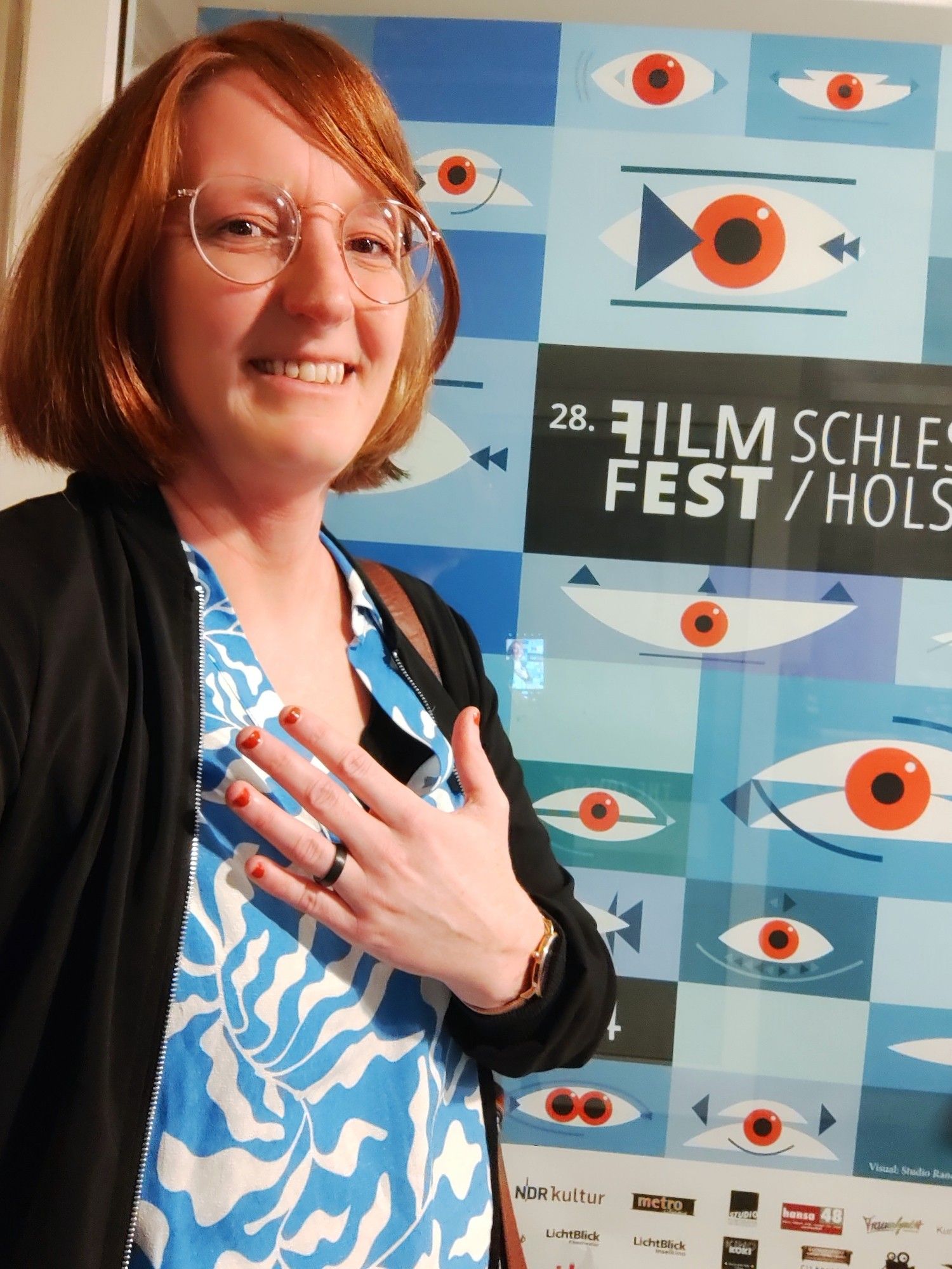 Sarah vor einem Festivalplakat - mein blau-weißes Shirt und dunkle Jacke korrespondieren perfekt mit den Blautönen des Plakats. Ich halte meine Hand mit roten Fingernägeln hoch, die genau den roten Augen im Plakat entsprechen.