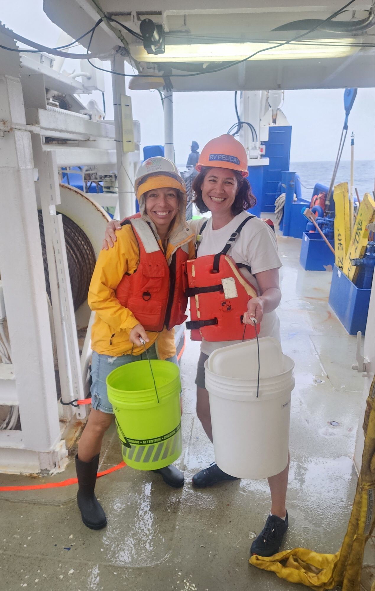 Chris Kellogg and Sarah Tweedt in hard hats and safety vests on ship’s deck in bad weather,  looking like they are about to go trick-or-treating for samples with buckets