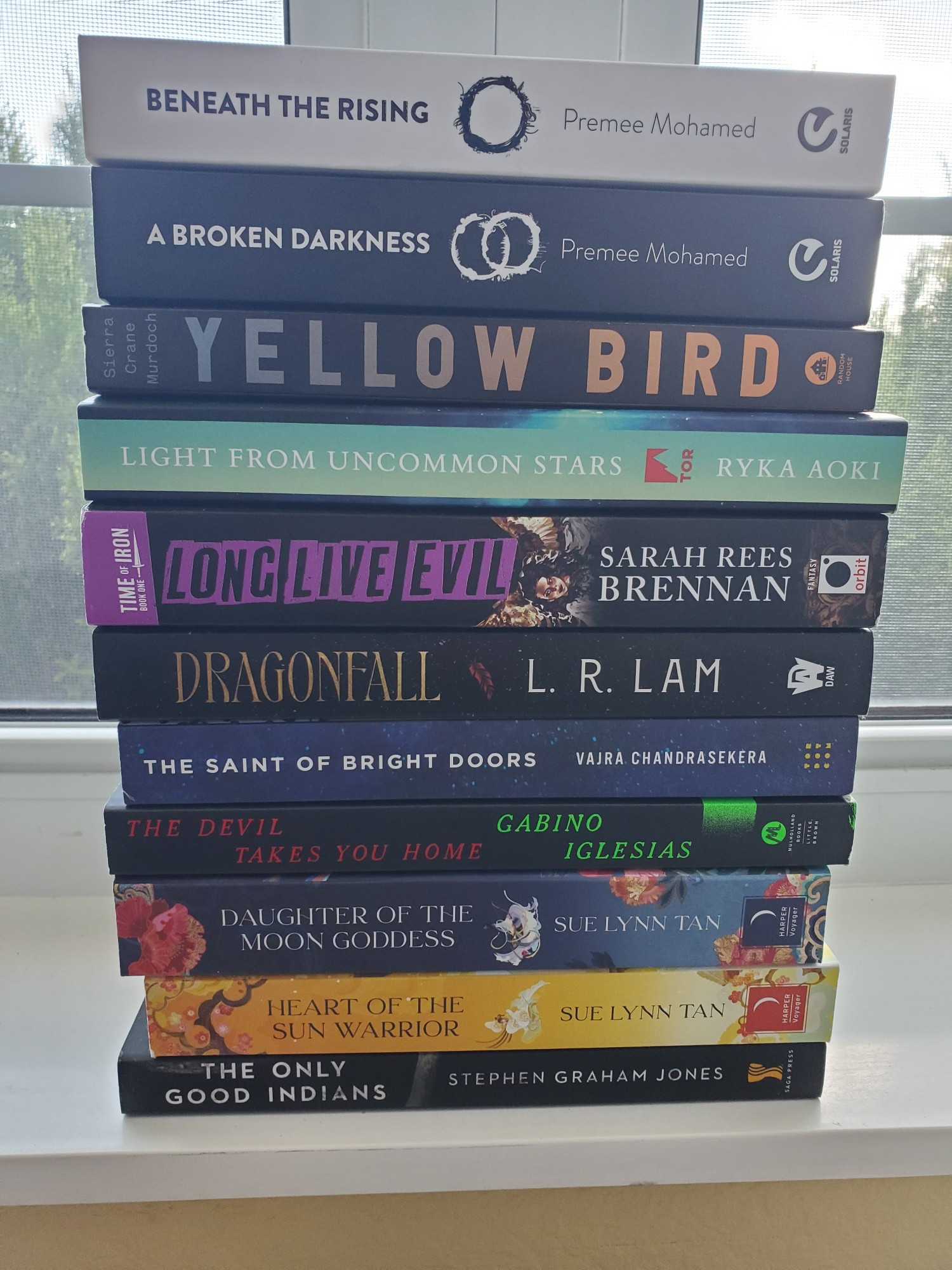 A stack of trade paperback books, top to bottom: Beneath the Rising by Premee Mohamed, A Broken Darkness by Premee Mohamed, Yellow Bird by Sierra Crane Murdoch, Light From Uncommon Stars by Ryka Aoki, Long Live Evil by Sarah Rees Brennan, Dragonfall by L.R. Lam, The Saint of Bright Doors by Vajra Chandrasekera, The Devil Takes You Home by Gabino Iglesias, Daughter of the Moon Goddess by Sue Lynn Tan, Heart of the Sun Warrior by Sue Lynn Tan, and The Only Good Indians by Stephen Graham Jones.