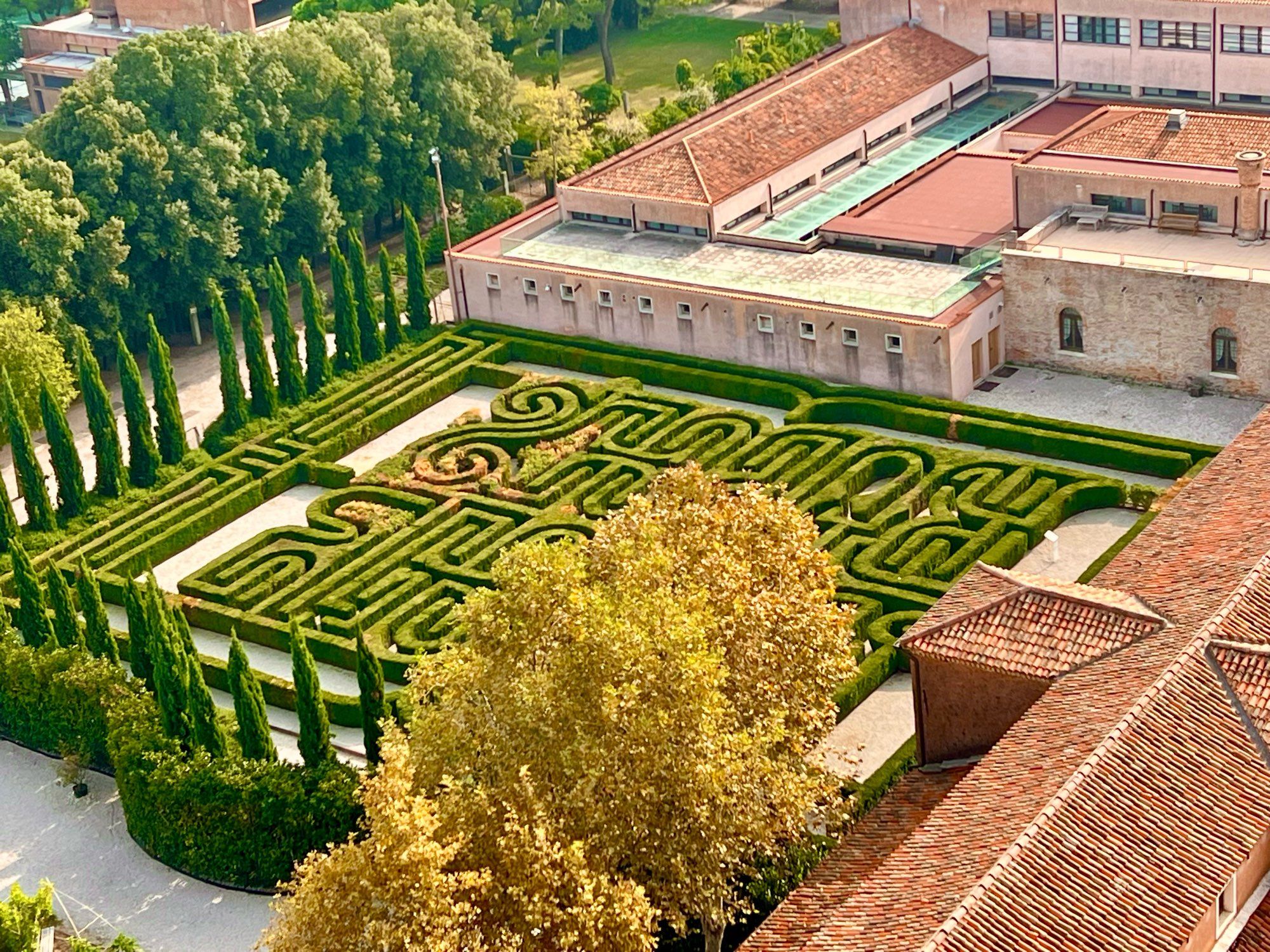 Blick von oben auf ein Garten- Labyrinth. Dahinter rote Gebäude.