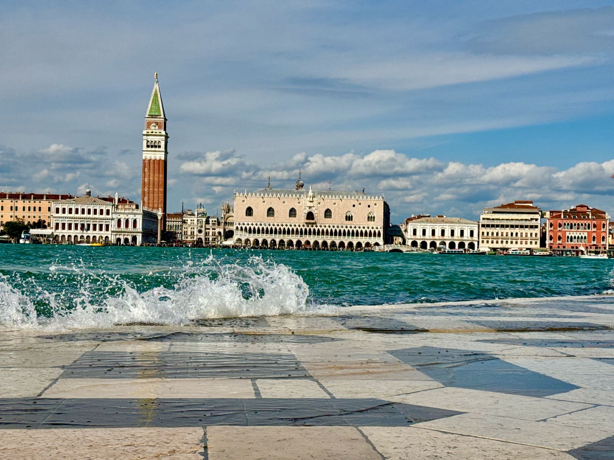 Das Wasser schwappt auf die Promenade, kleine Wellen brechen sich. Im Hintergrund der Campanile des Markusdoms und andere Gebäude.