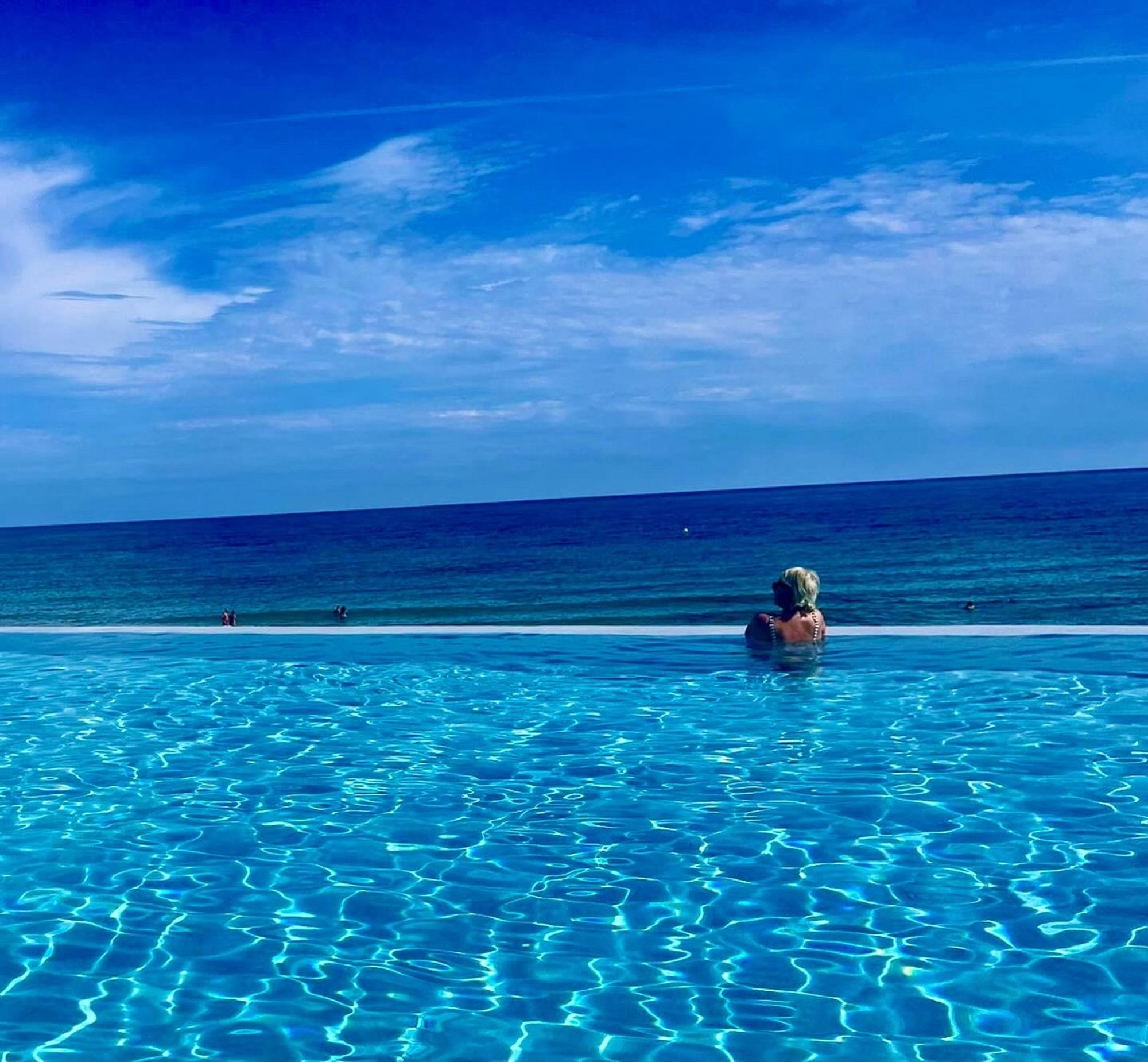 Blauer Himmel, blaues Meer, blauer Pool ineinander übergehend. Eine Frau schaut aufs Meer, im Pool stehend.