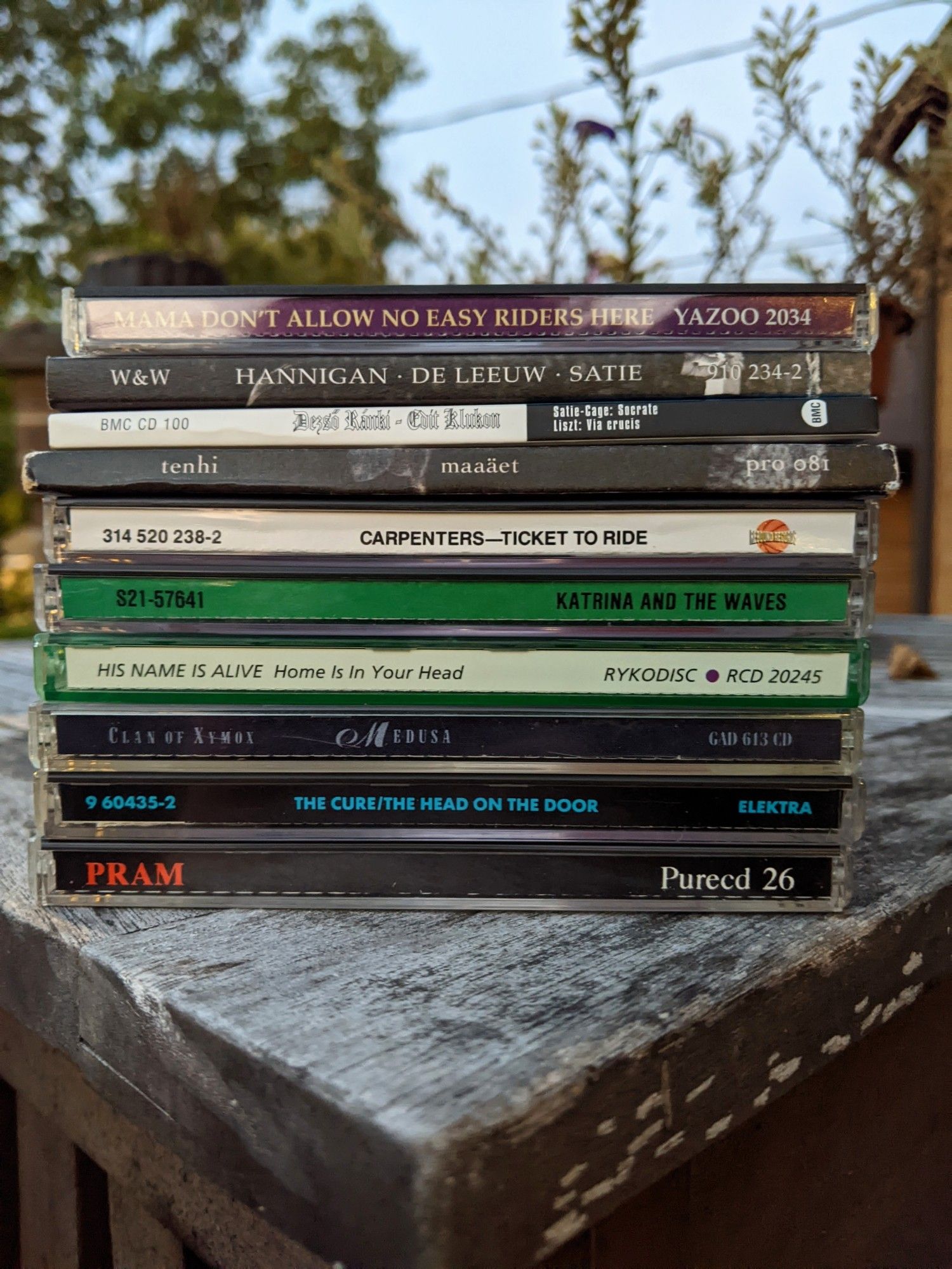 Stacked CD playlist on weathered wooden patio table, set at an approximately equilateral angle to make a triangle with the corner of the table. Pre-dawn light, digitally brightened.

In order played, bottom to top:

Pram THE STARS ARE SO BIG, THE EARTH IS SO SMALL... STAY AS YOU ARE
The Cure THE HEAD ON THE DOOR
Clan of Xymox MEDUSA
His Name is Alive HOME IS IN YOUR HEAD
Katrina and The Waves S/T
Carpenters TICKET TO RIDE
Tenhi MAAAËT
Satie:Cage SOCRATE, Liszt VIA CRUCIS (Dezső Ránki - Edit Klukon, pianos)
Satie SOCRATE (Barbara Hannigan - Reinbert de Leeuw) 
Various MAMA DON'T ALLOW NO EASY RIDERS HERE